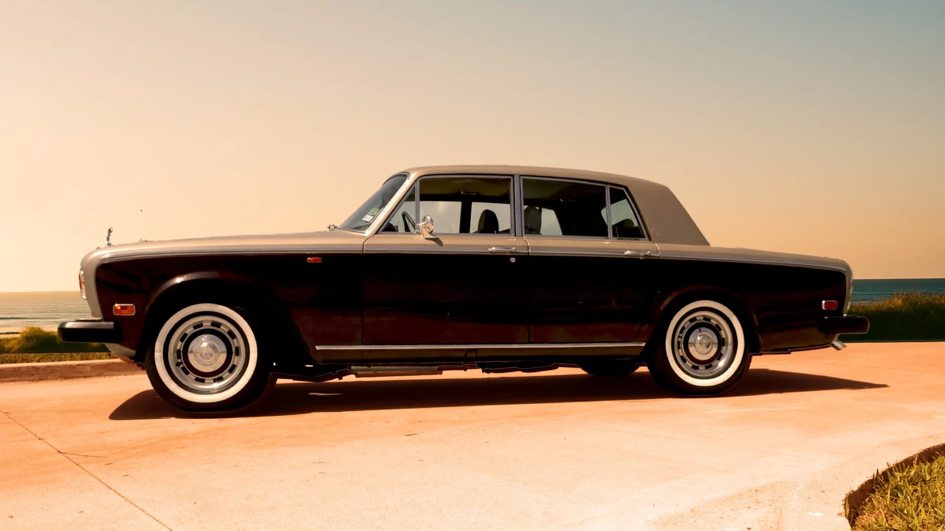A vintage black and silver luxury car parked on a concrete surface near the coast at sunset