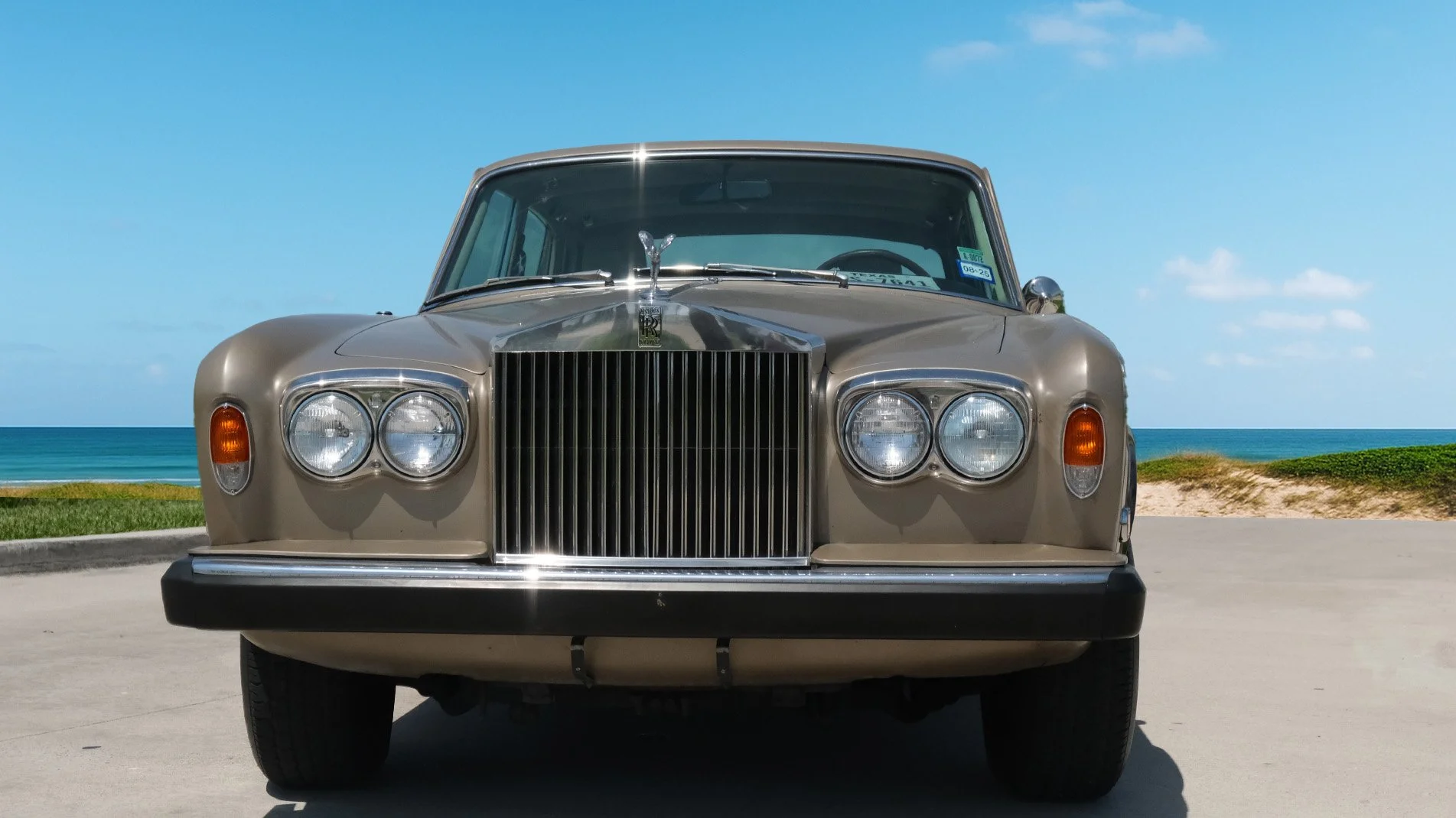 Front view of a vintage beige Rolls-Royce car parked near a beach with ocean and sky in the background.