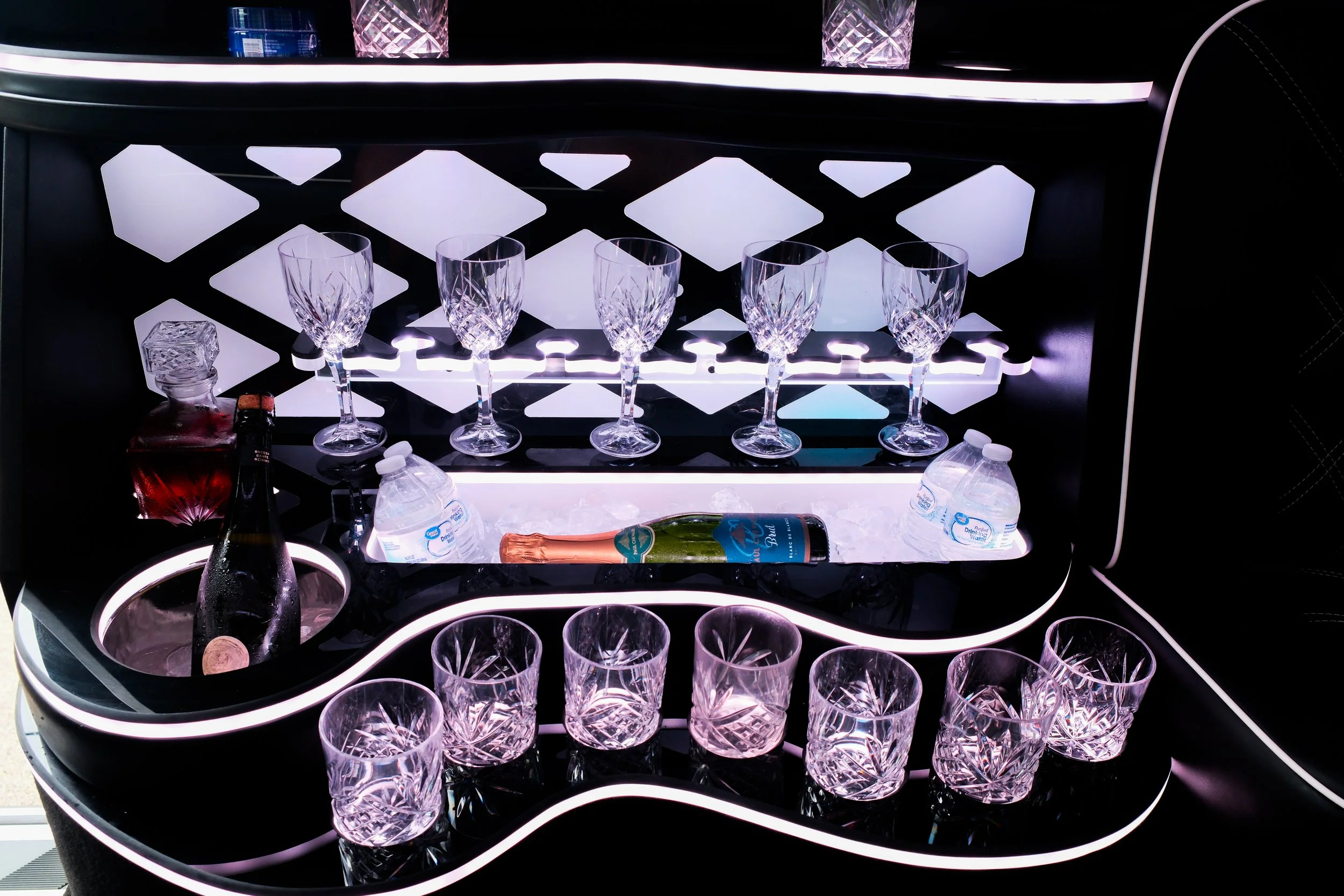 A stylish bar cart with illuminated shelves holding crystal glasses, a bottle of champagne, bottled water, and a bottle of other liquor, all against a dark background with modern lighting accents.
