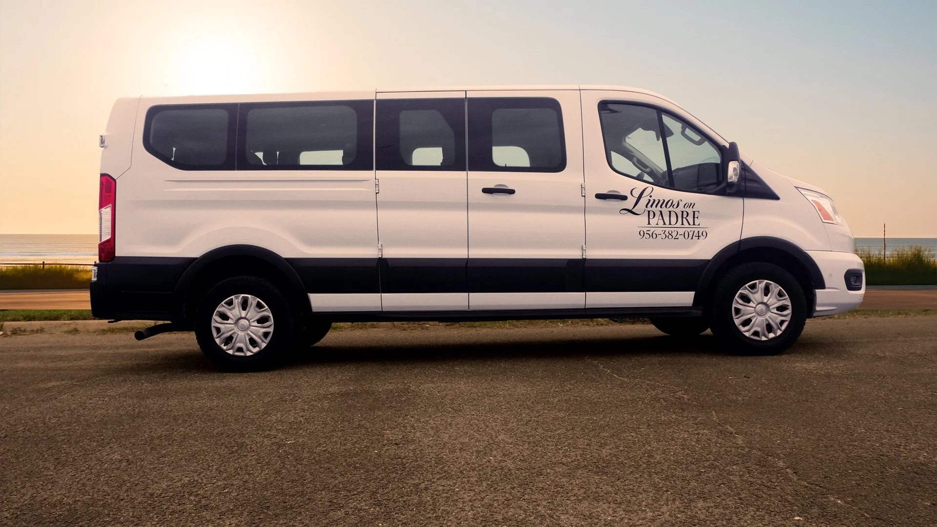 A white Passenger van parked on a beachside parking lot at sunset with the ocean and a clear sky in the background. The vehicle has a logo and phone number on its door.