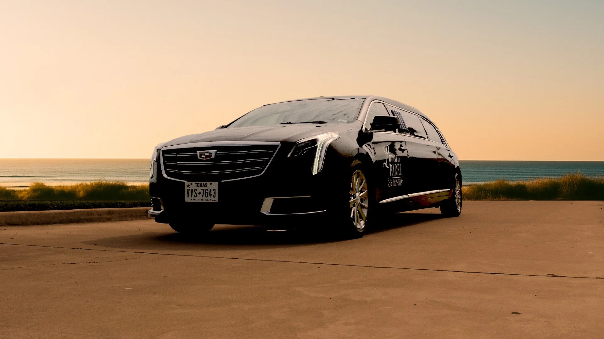 Black Cadillac limousine with Texas license plate parked near a beach at sunset, with the ocean and sky in the background.