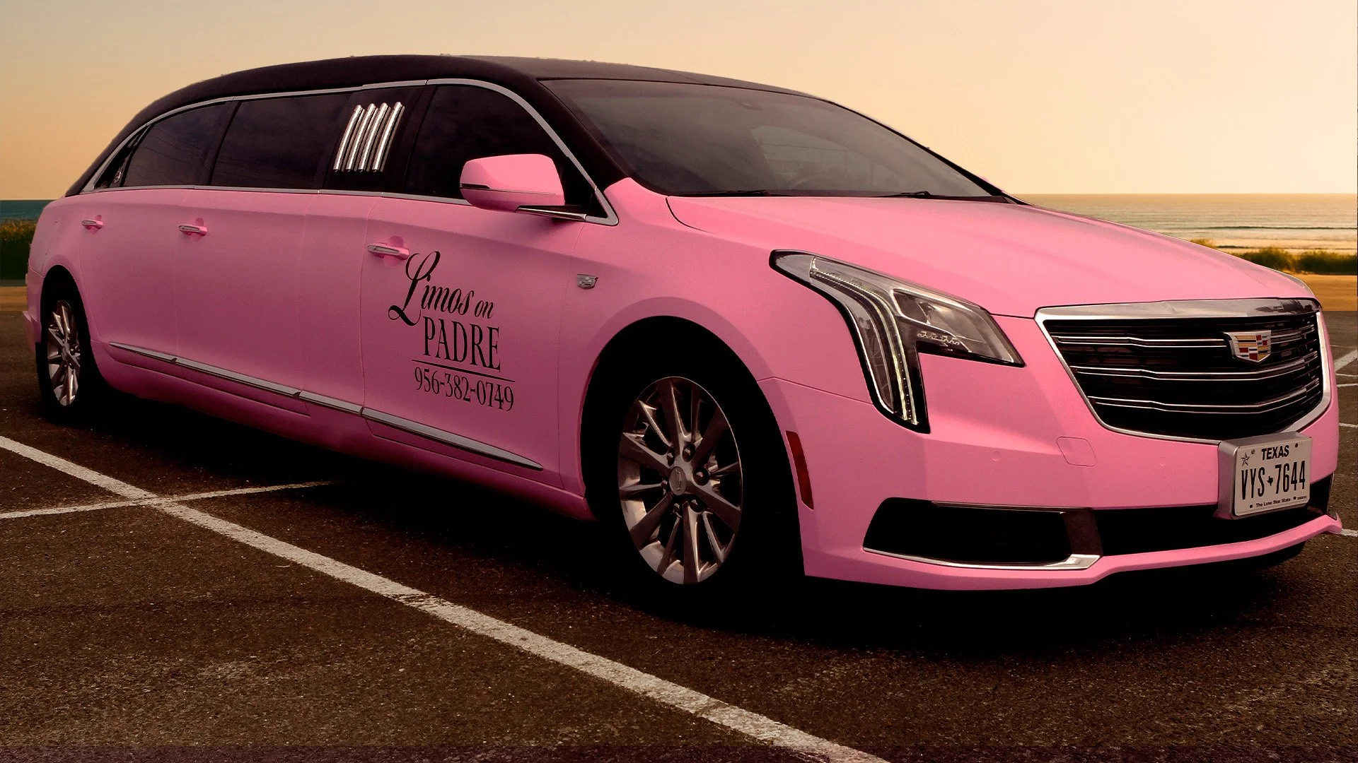Pink Cadillac with lettering that reads "Limos en PADRE" and a phone number, parked near the ocean at sunset.