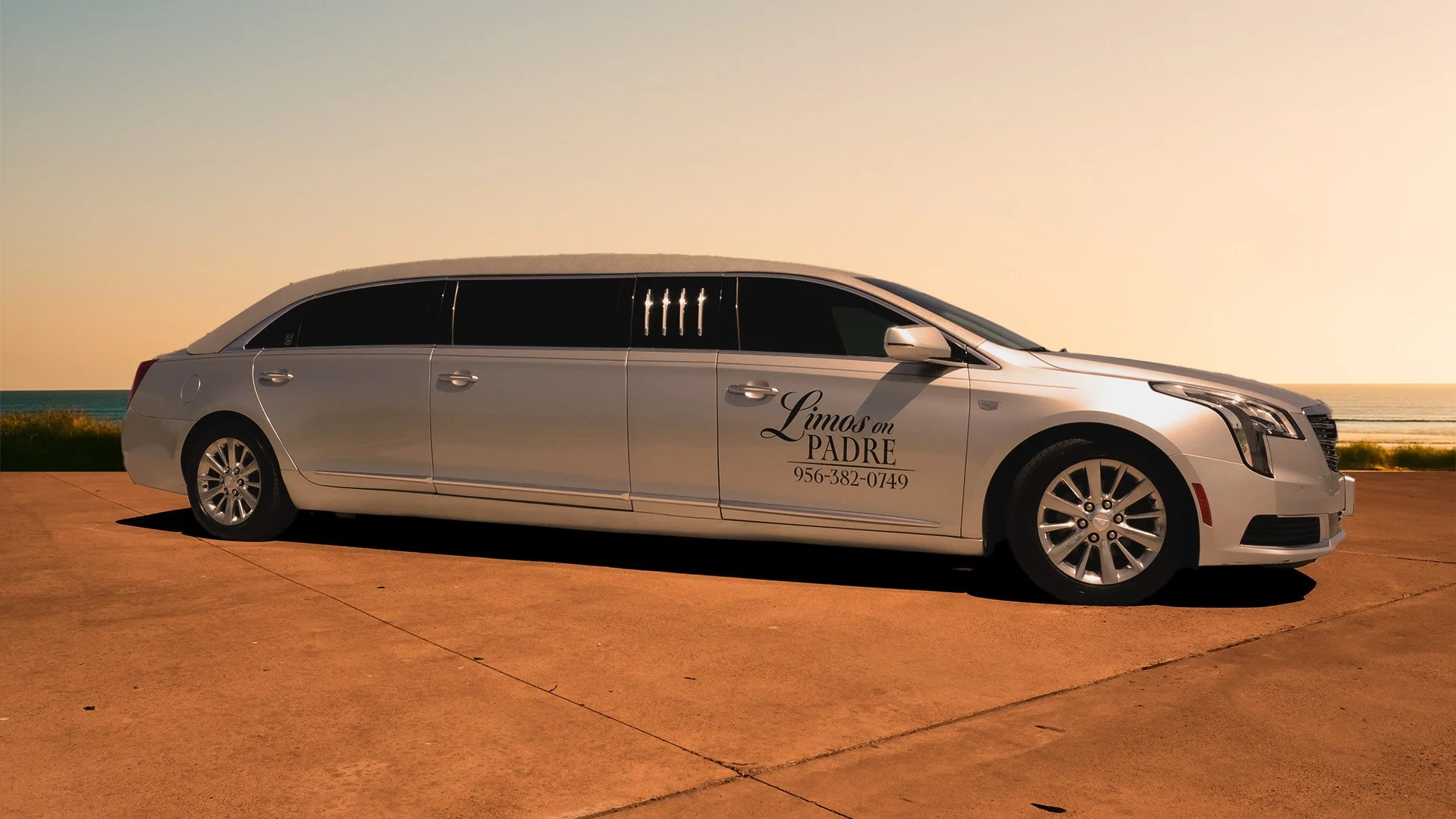A silver stretch limousine parked on a concrete surface with a seaside background during sunset. The limousine has black tint windows and decal text that reads, "Limos on PADRE" with a phone number.