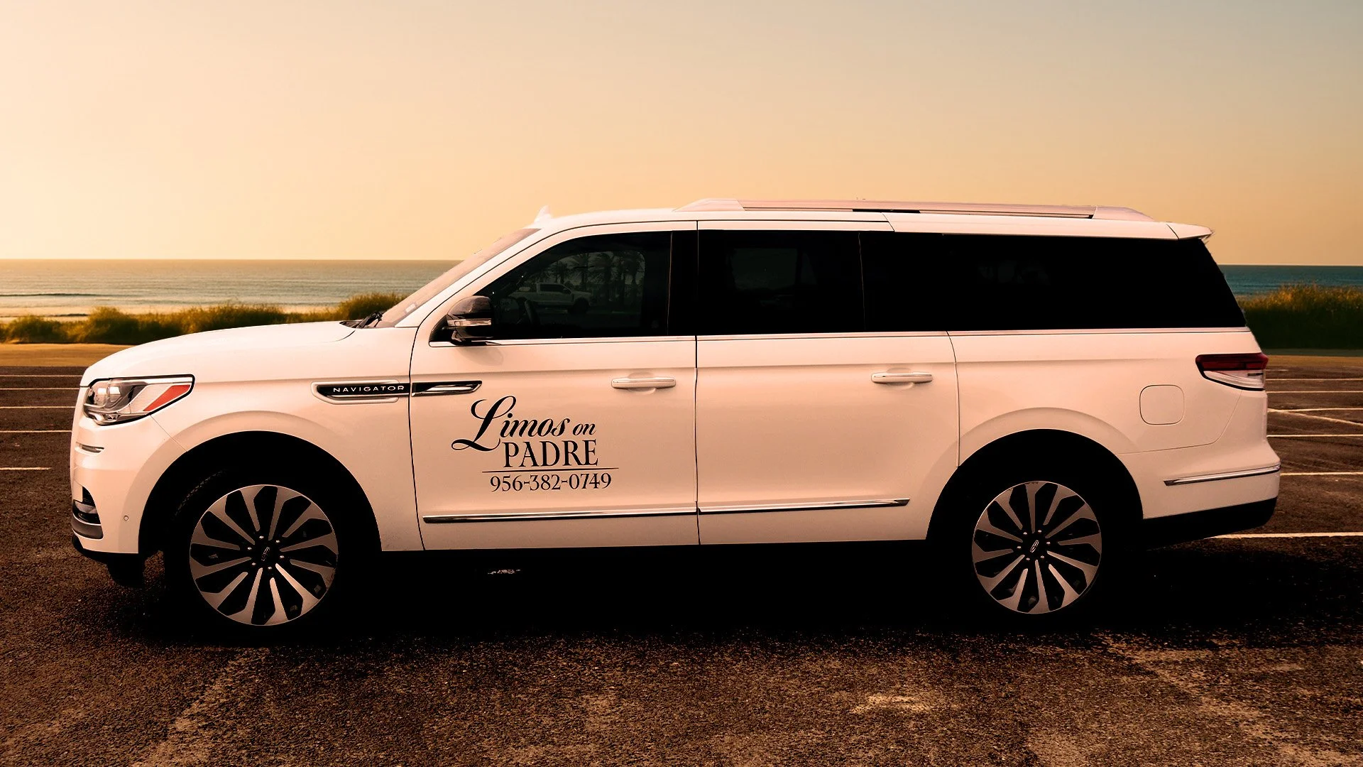 A white limousine parked on a beachside parking lot at sunset with the ocean and a clear sky in the background. The vehicle has a logo and phone number on its door.