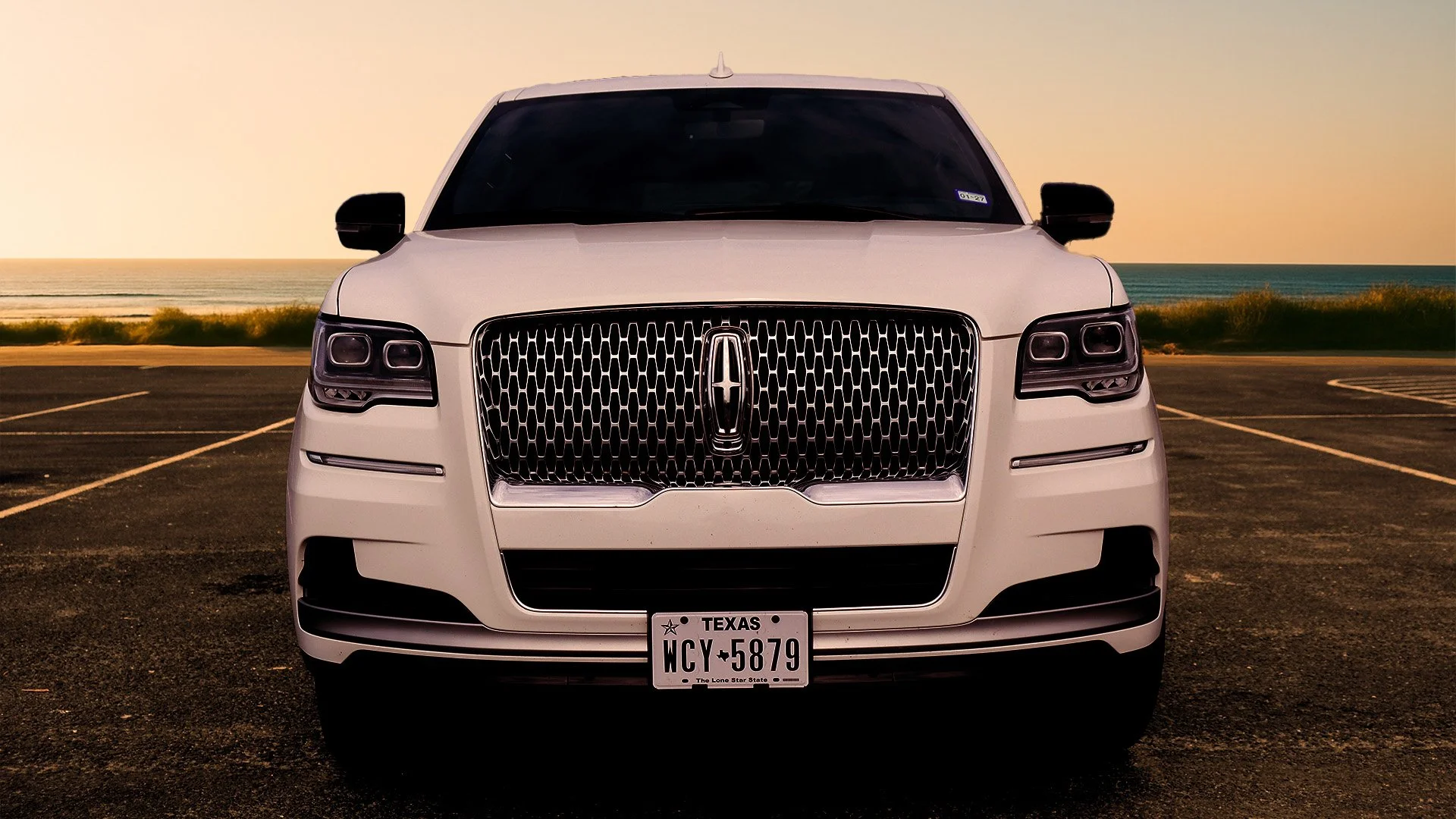 front view of a white luxury SUV parked near the beach at sunset with an ocean background