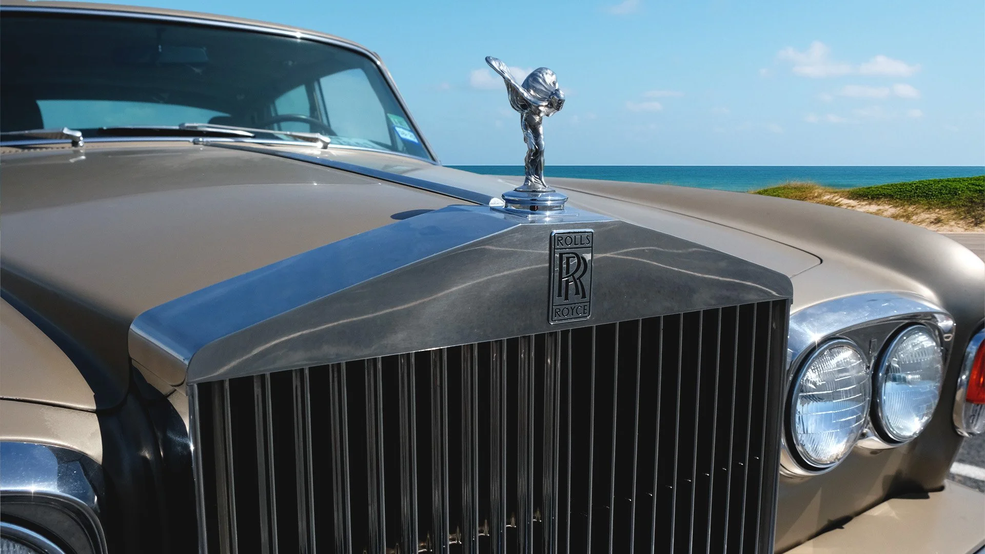 Close-up of a vintage Rolls-Royce car with a silver hood ornament, parked near the beach under a blue sky, showing the grille and front headlights.