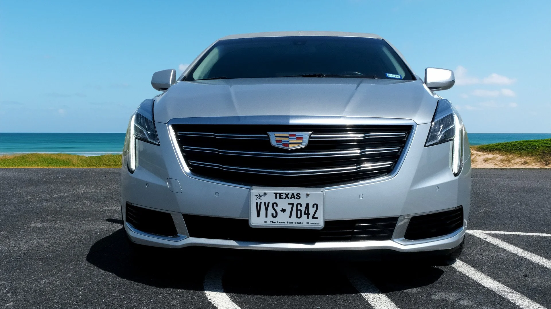 Front view of a silver Cadillac car parked on a blacktop surface near a coast with sandy beach and ocean in the background.