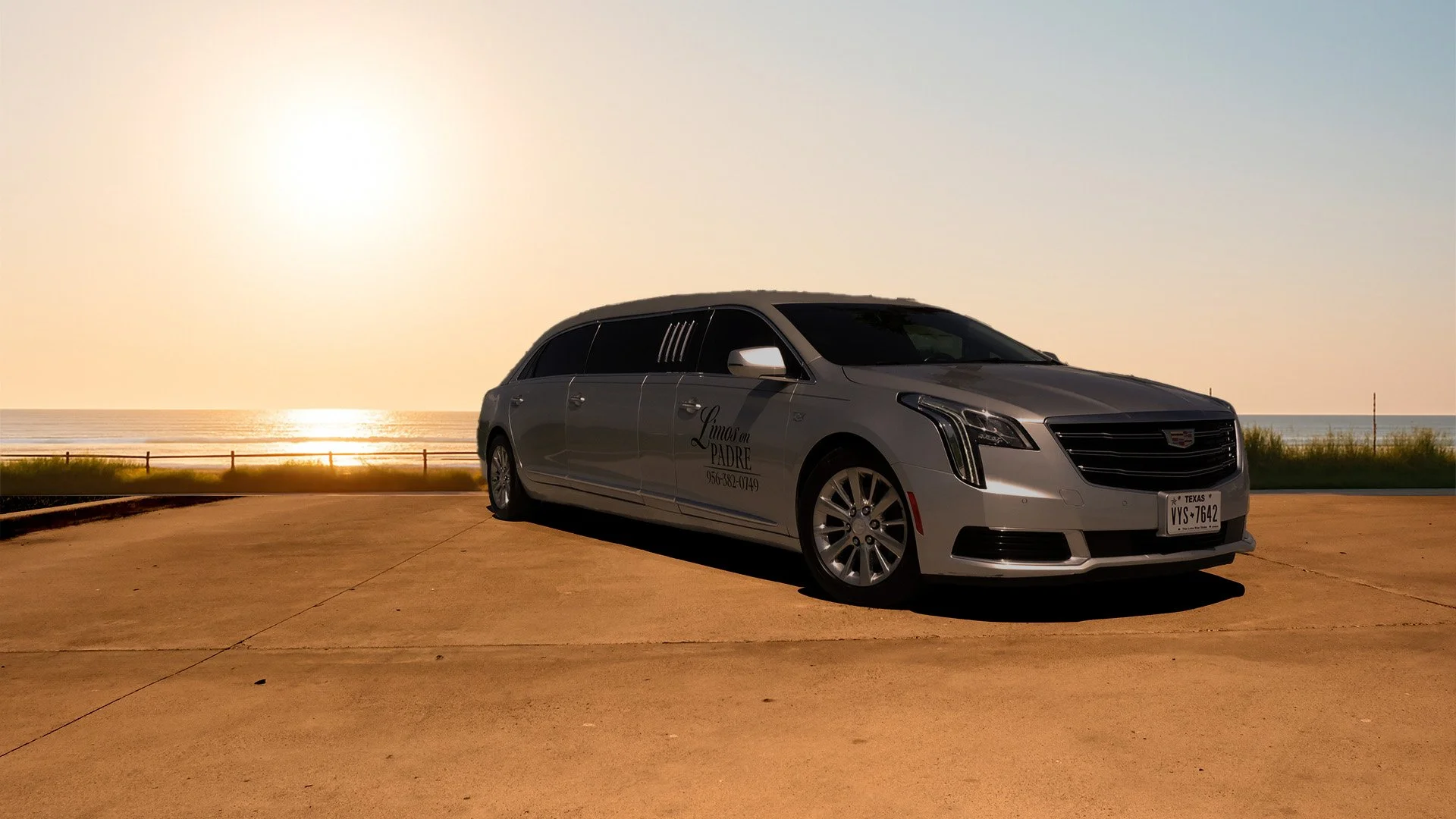 A silver Cadillac limousine parked on a beachside pavement with the ocean and sunset in the background.