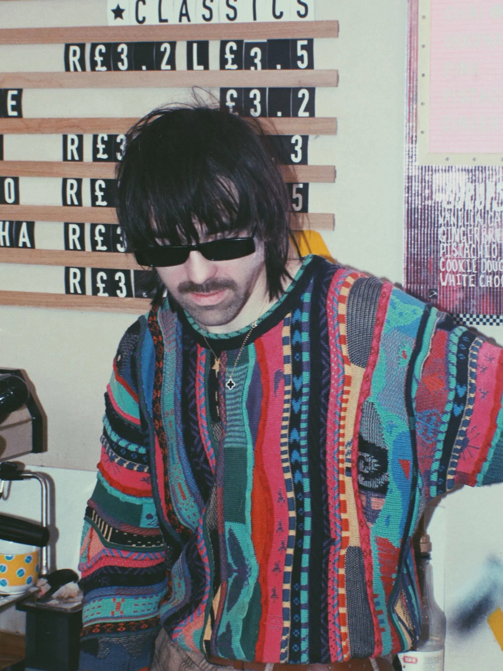 A person wearing black sunglasses and a colorful, patterned sweater with vertical stripes and various shapes, standing in front of a wall with a price list and a poster.