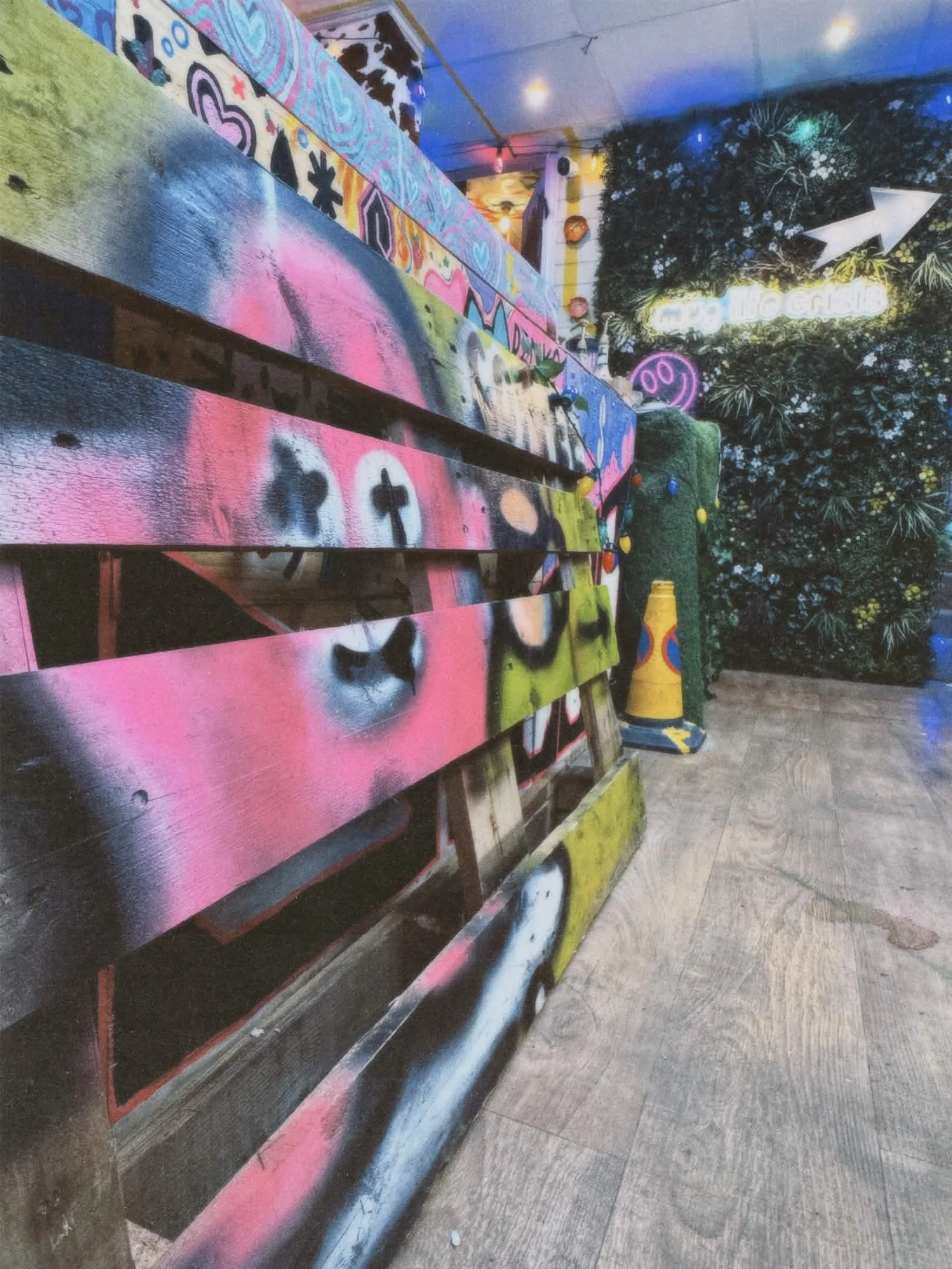 Colorful graffiti art on a wooden bench and wall, neon signs, and a dense green plant wall in a trendy indoor space.