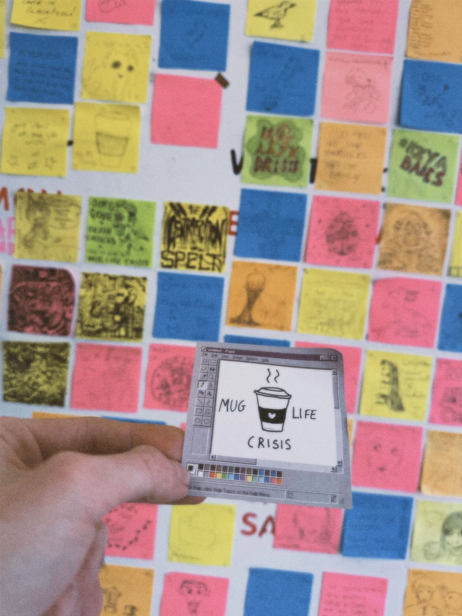 A hand holding a small drawing of a coffee cup labeled 'MUG LIFE CRISIS' in front of a colorful collage wall with sticky notes and drawings.