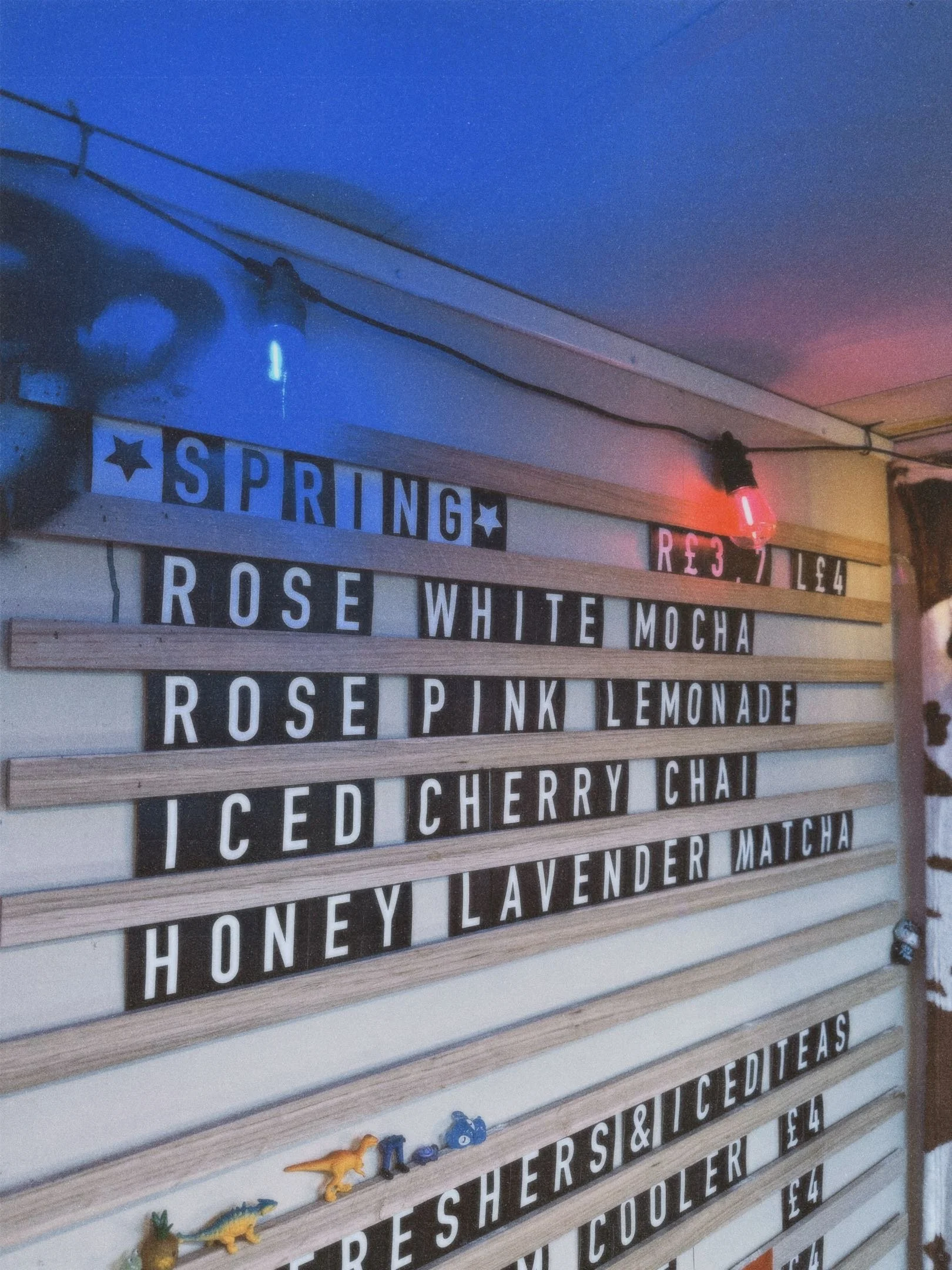 A menu board listing various drinks such as rose white mocha, rose pink lemonade, iced cherry chai, honey lavender matcha, and others, with a neon light and some small decorative toys on the bottom shelf.