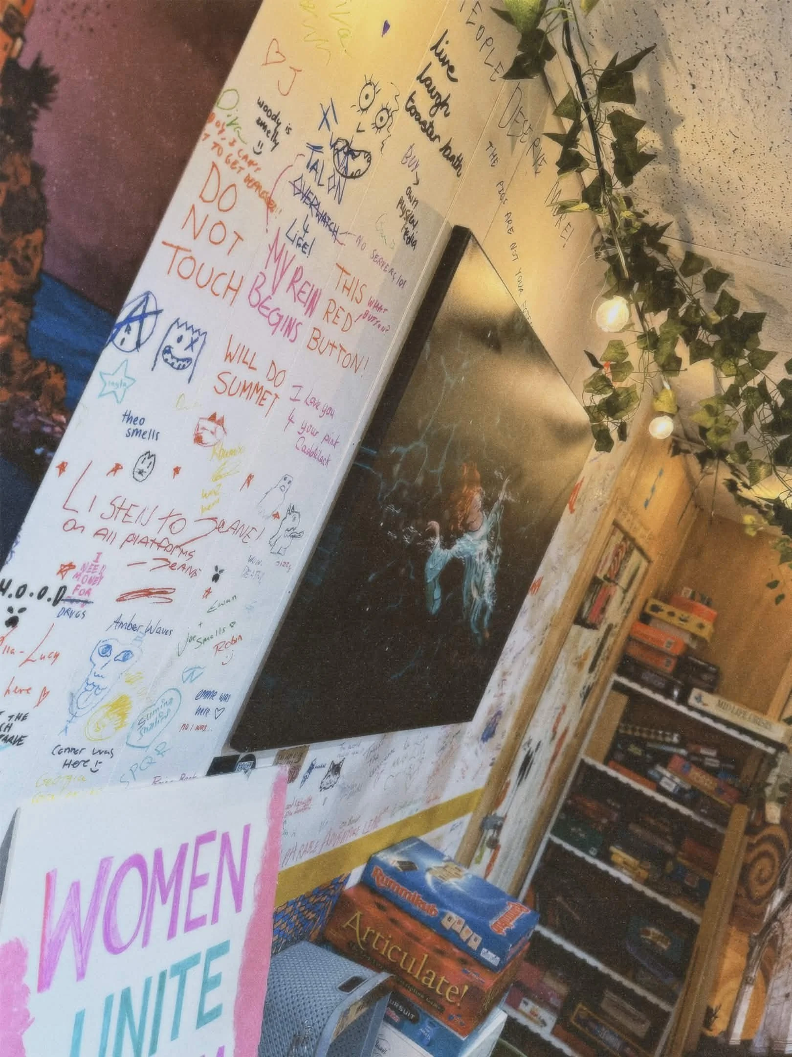 A wall filled with colorful handwritten notes, doodles, and messages, with a black artwork featuring an abstract figure in the center. There is also greenery with string lights hanging overhead.