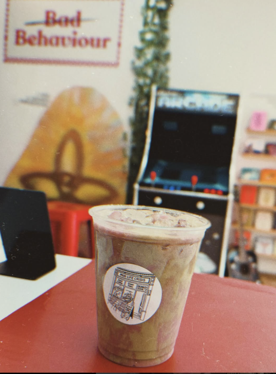 Blueberry Açaí Oat Shaken Matcha served iced in a clear cup with Mug Life Crisis café logo, placed on a red counter with arcade machine and retro décor in the background