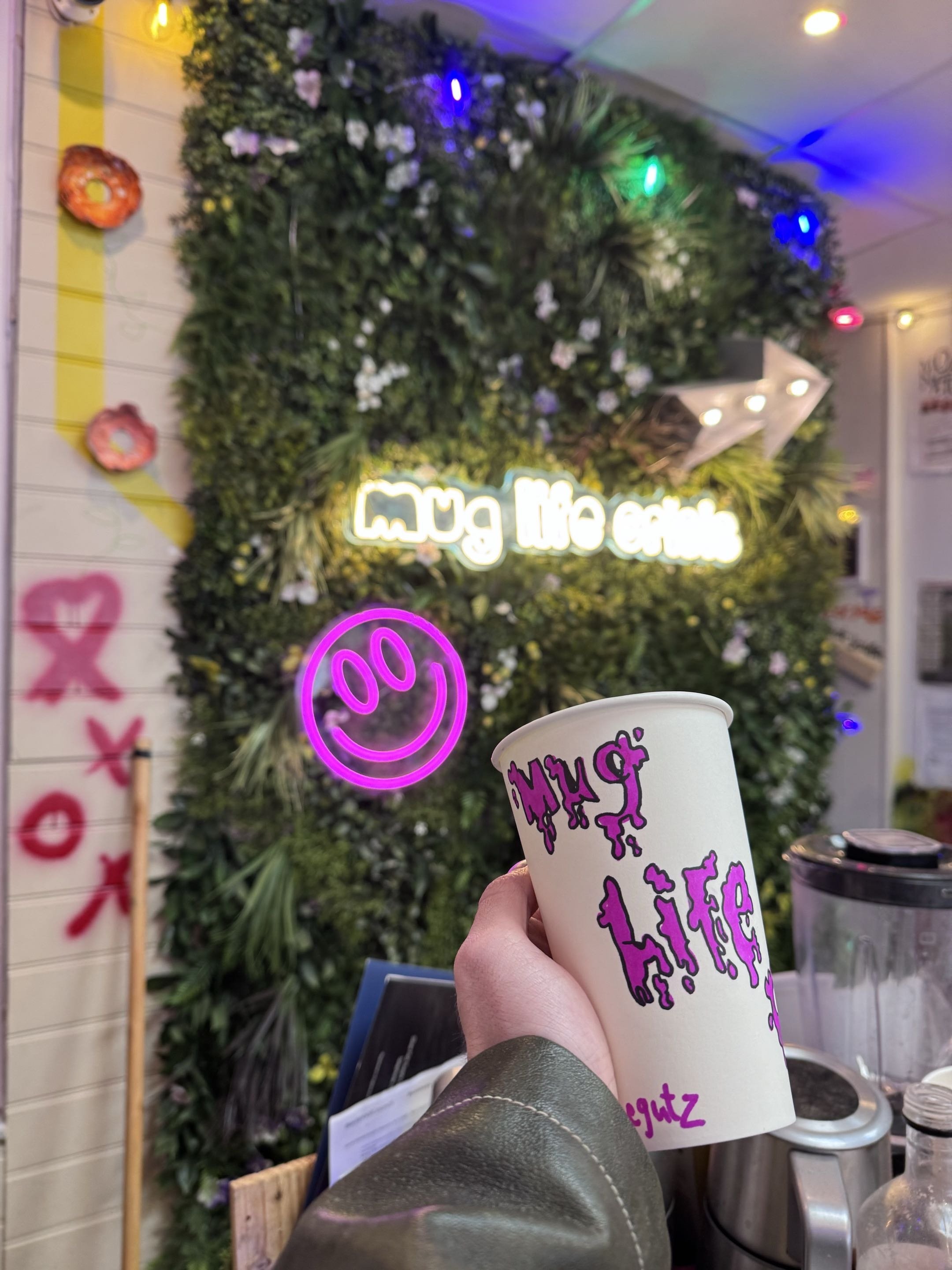 A person holding a white paper cup with purple text on it in front of a decorated wall with a neon sign, a smiley face, and colorful string lights, inside a cafe or shop.