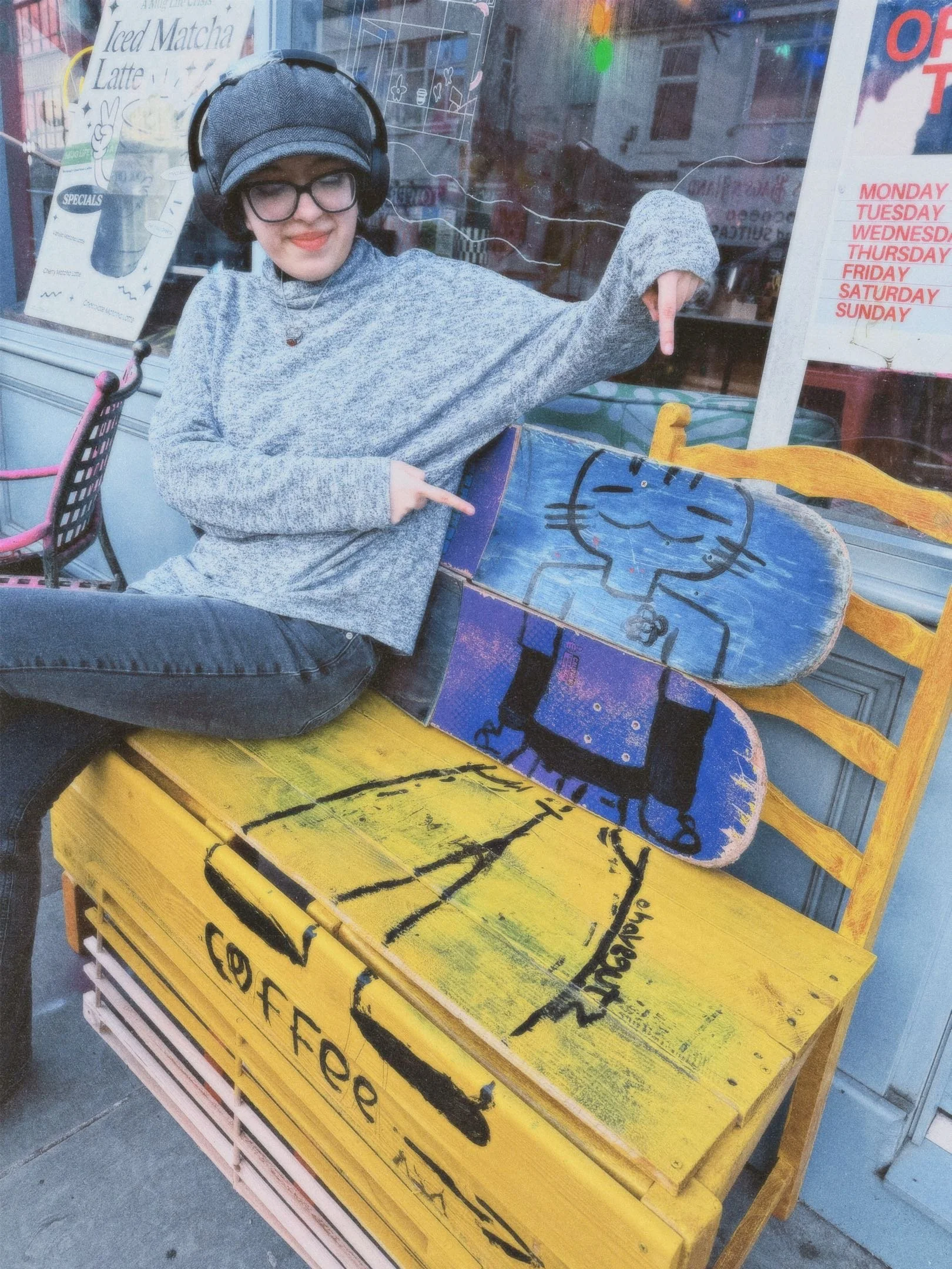 A person wearing glasses and a gray sweater, sitting outdoors at a yellow table with a skateboard featuring a cartoon cat drawing, pointing at the skateboard. There are colorful chairs and a store window in the background with signs and menu boards.