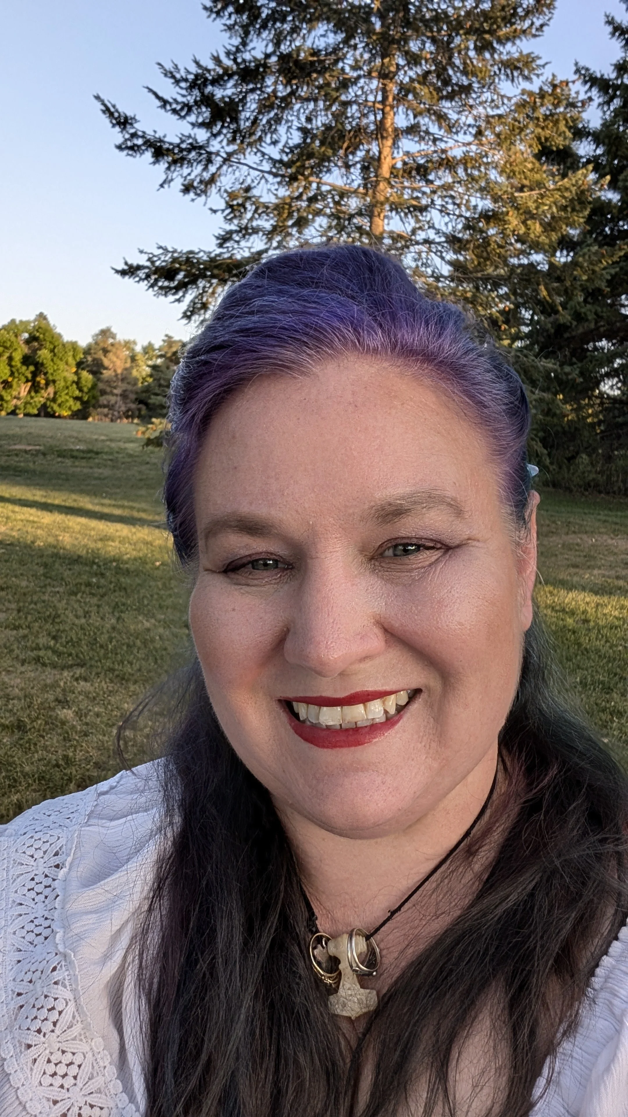 A woman with purple and black hair smiling outdoors, wearing a white lace top and a necklace with a carved pendant, with trees and a grassy field in the background.