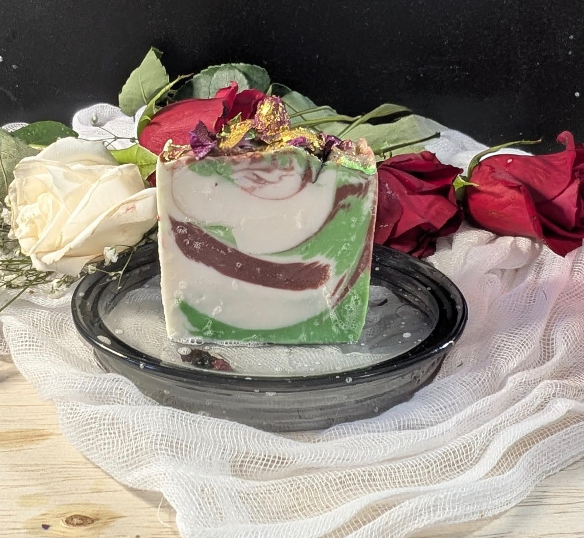 A decorated soap bar with swirling green, white, and red colors, topped with floral and shiny embellishments, placed on a black glass plate with roses in the background.