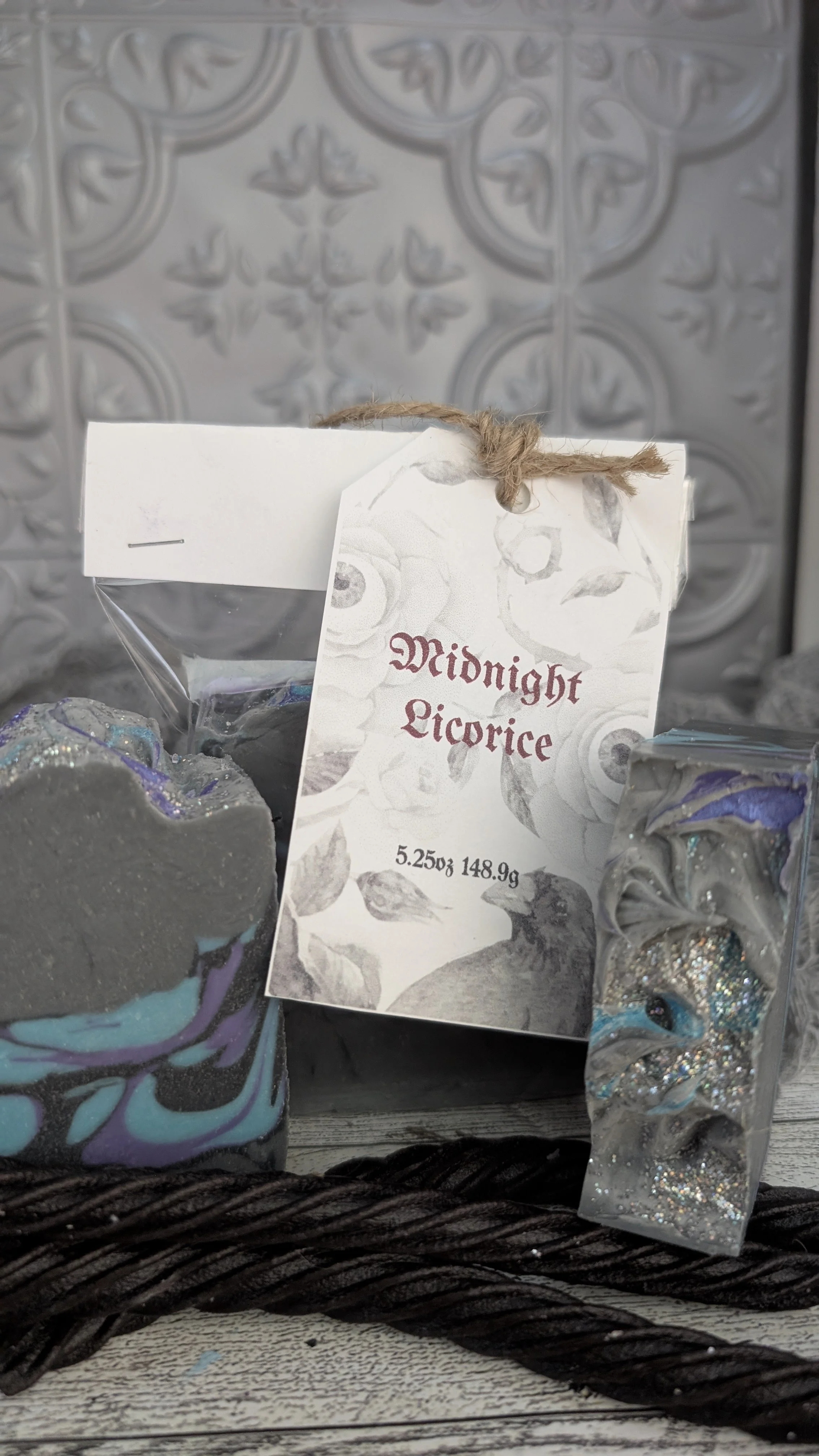 A decorative soap with swirling purple, teal, and black colors, next to a soap with glitter and swirled silver, with a label reading "Midnight Licorice," placed in front of a textured gray wall.