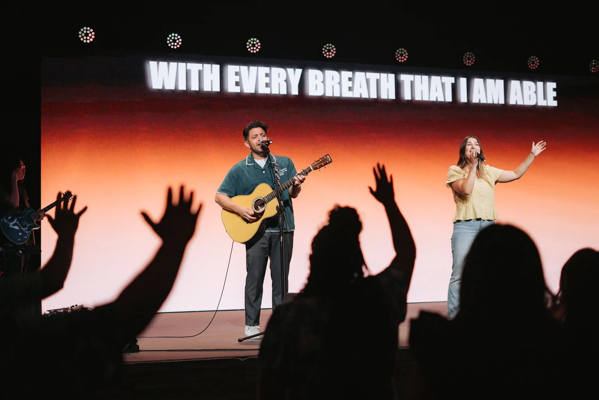 A band performs on stage with a large screen behind them displaying the lyrics 'WITH EVERY BREATH THAT I AM ABLE'. The audience has raised hands.