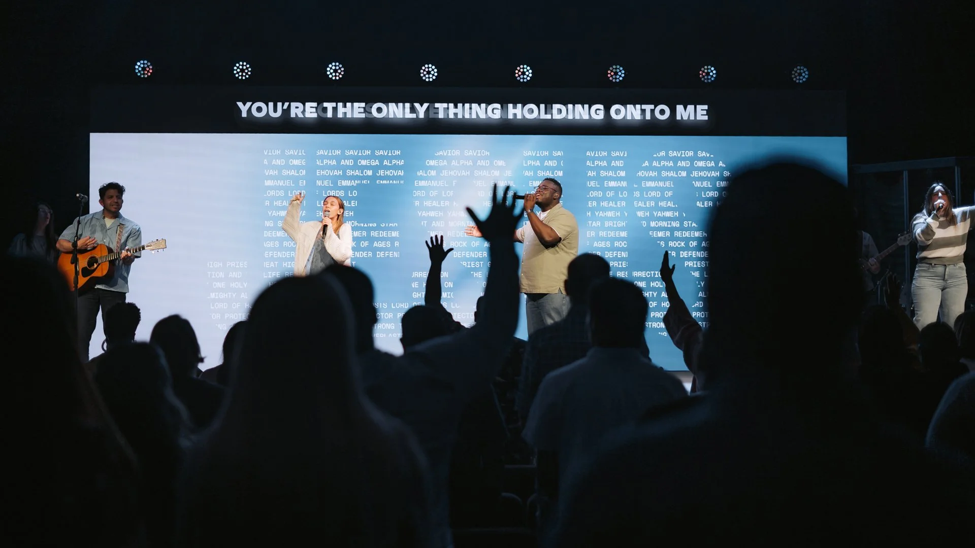 A group of musicians and singers performing on stage with a large screen behind them displaying song lyrics, titled "You're the only thing holding onto me." The audience is raising their hands.