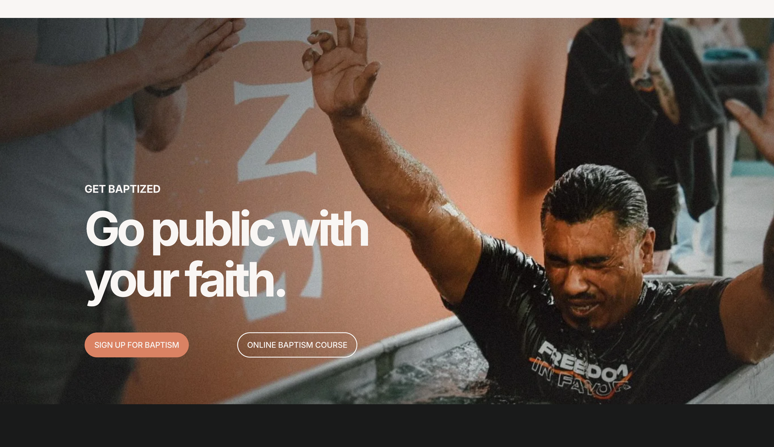 A man with slicked-back hair being baptized by immersion in a baptismal font, with his eyes closed and expression showing emotion, while a person nearby raises their hand and another person takes a photo. The background features a large 'Z' on an orange wall.