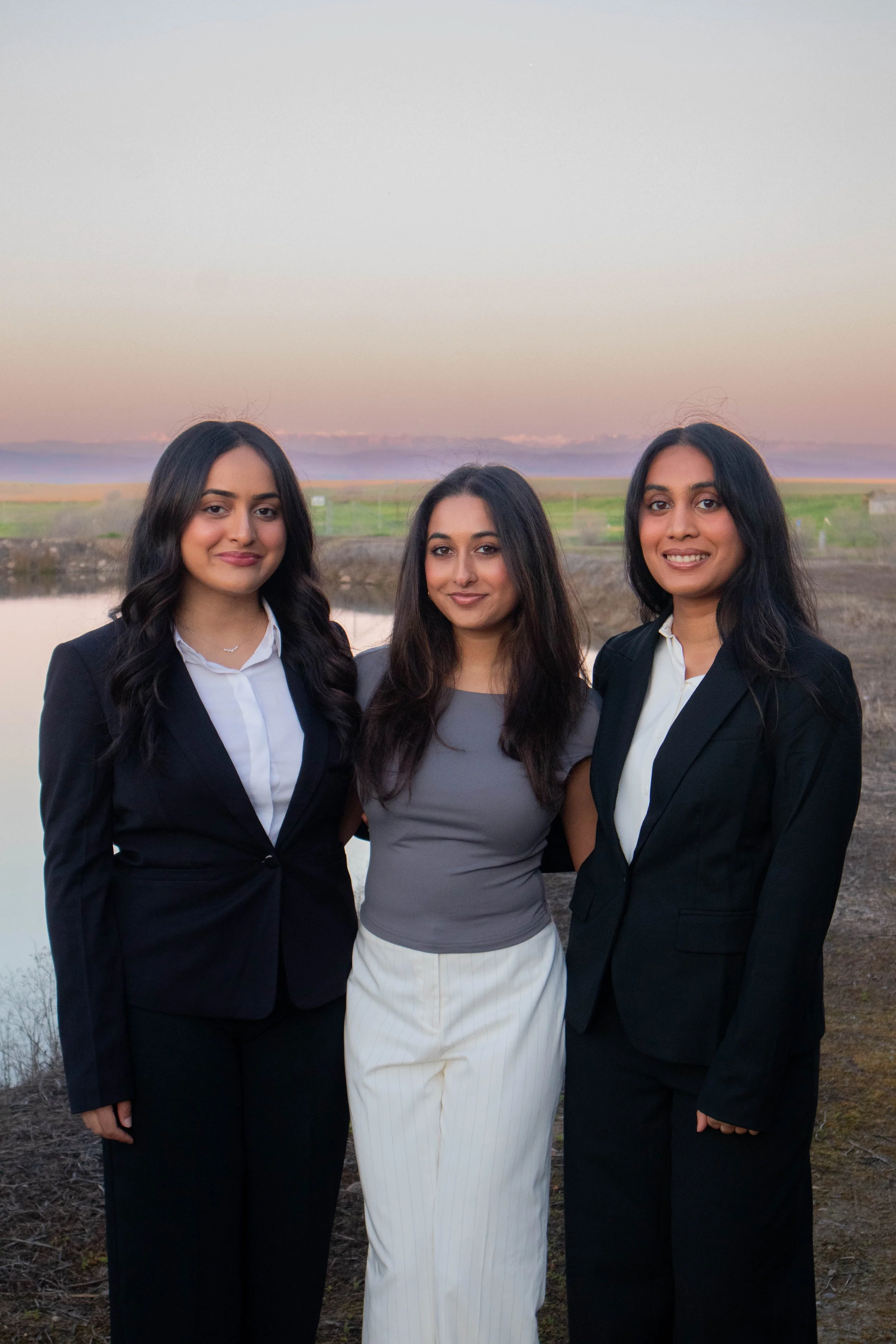 Three women posing outdoors in business attire with a field and sunset sky in the background.