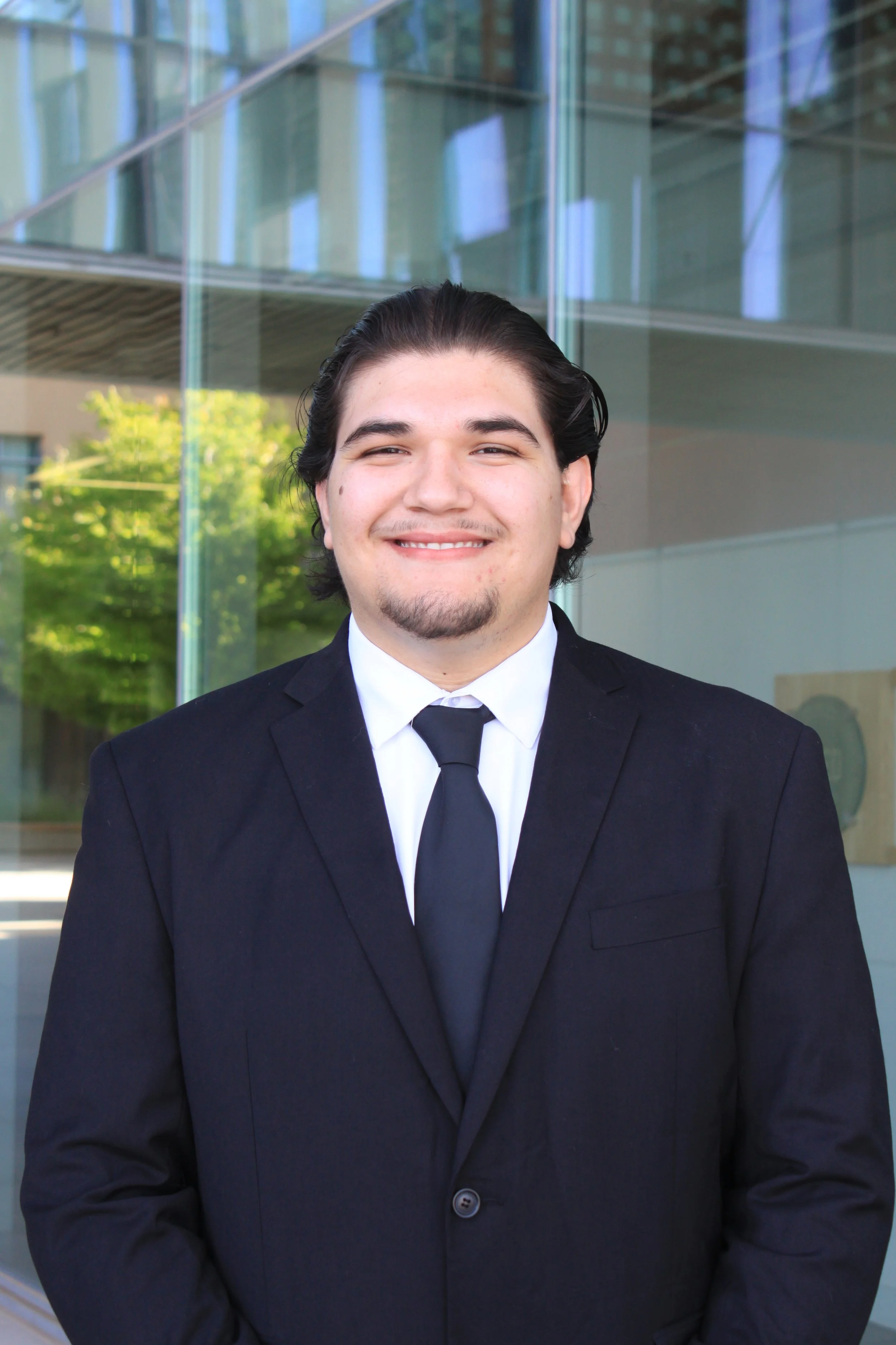 A person with long hair wearing a black blazer and white shirt stands in front of a modern glass building.