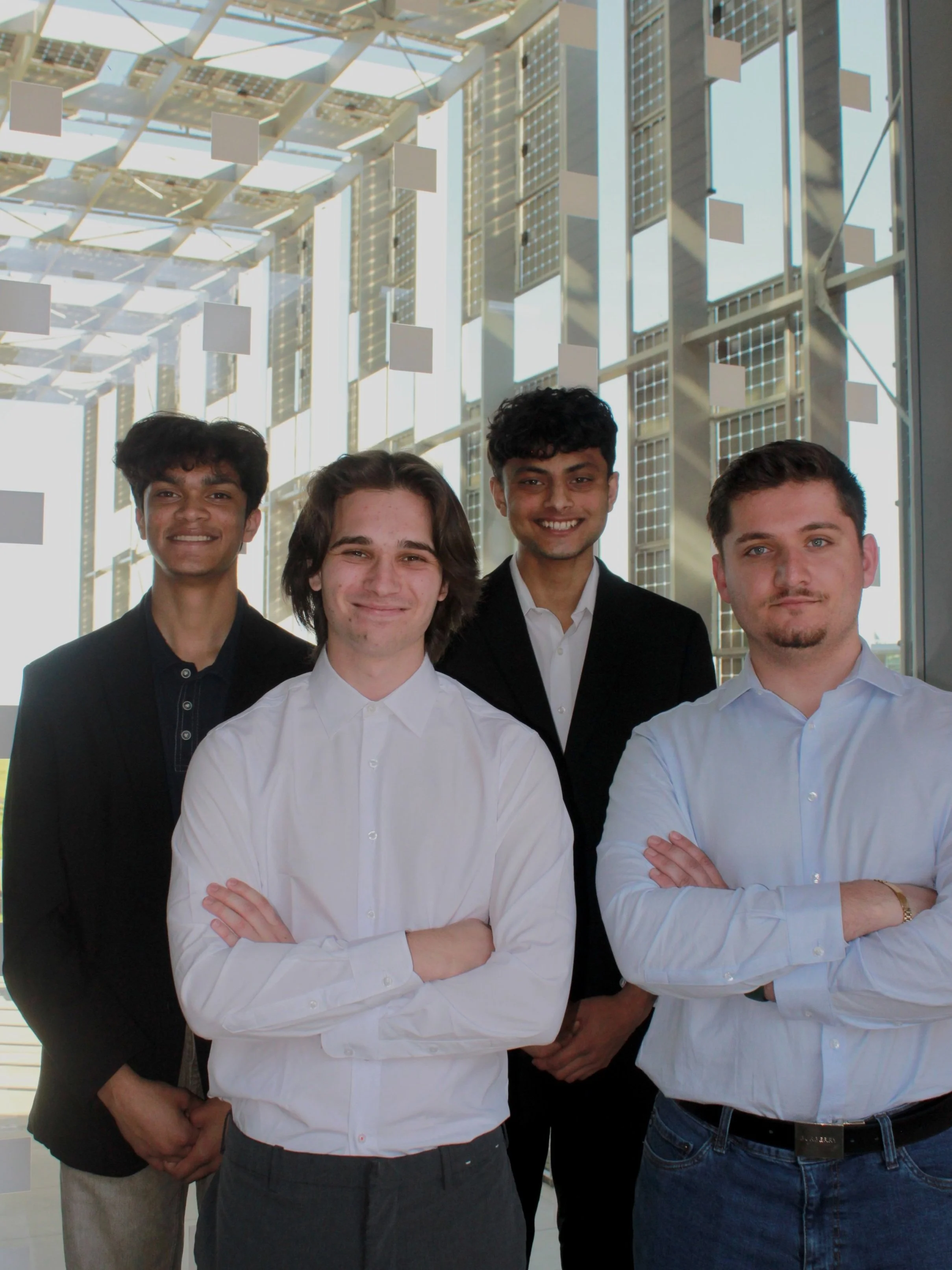 Group of four young men in business attire standing in a modern building with large glass windows and a backdrop of solar panels and architectural structures.