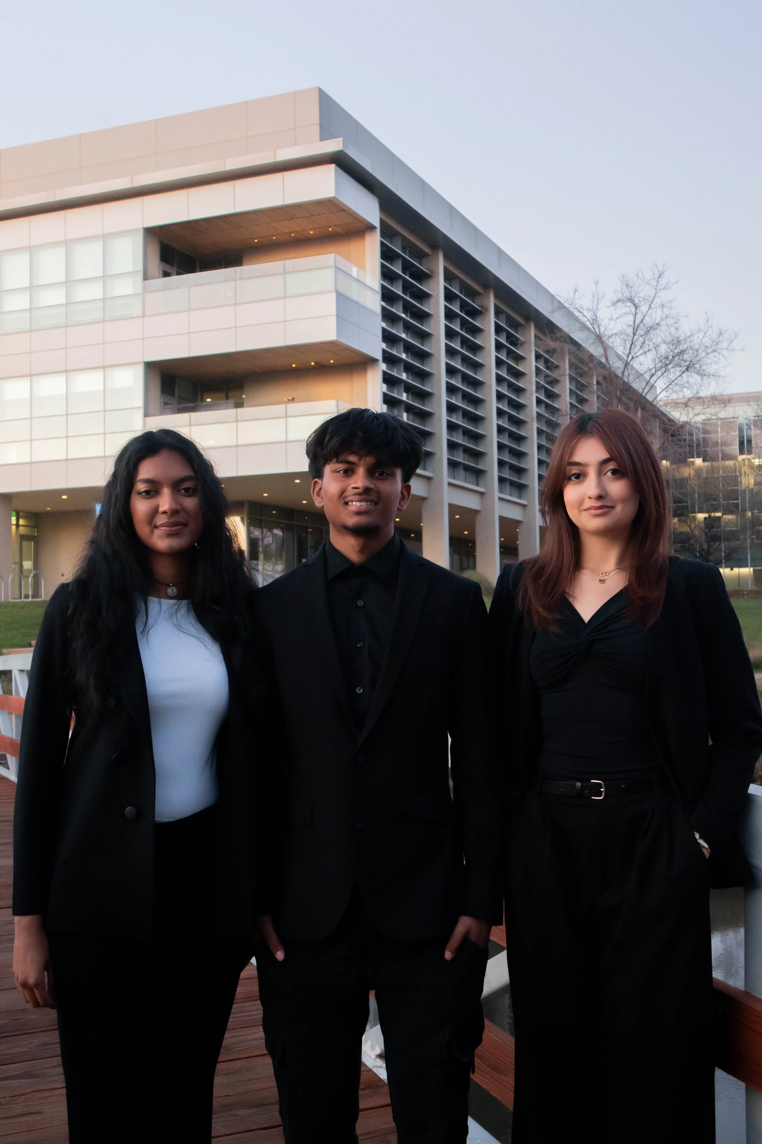 Three people in formal attire standing in front of a modern building, smiling at the camera.