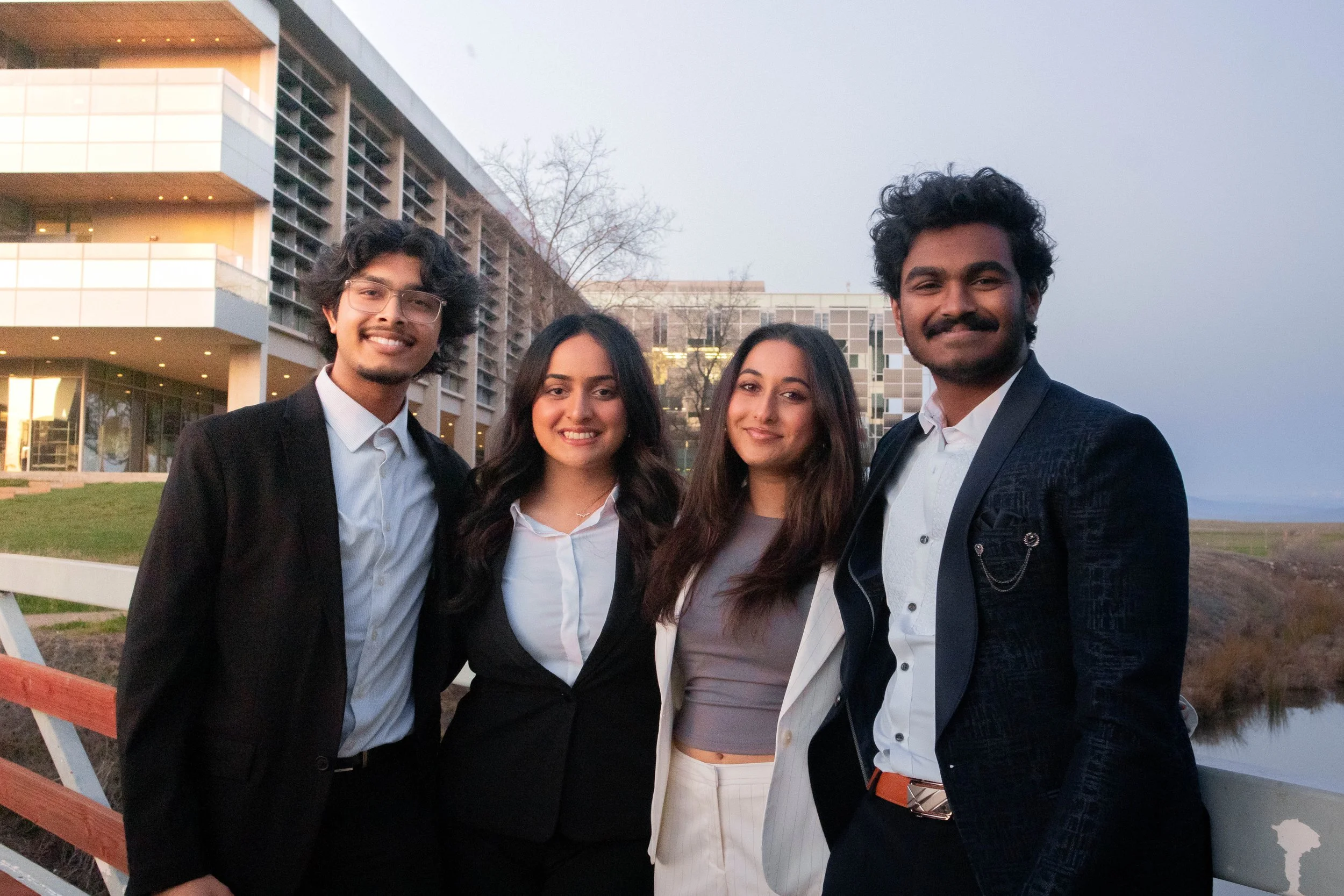 Group of four people in formal attire posing outdoors in front of a modern building.