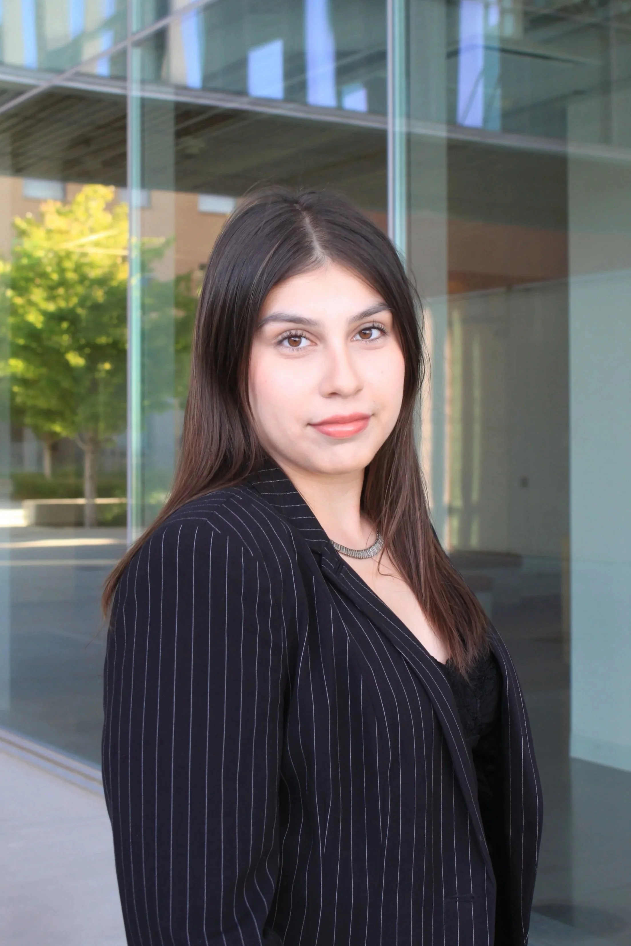 Young woman with shoulder-length brown hair, wearing a black pinstripe blazer and black top, standing outdoors in front of modern glass building.