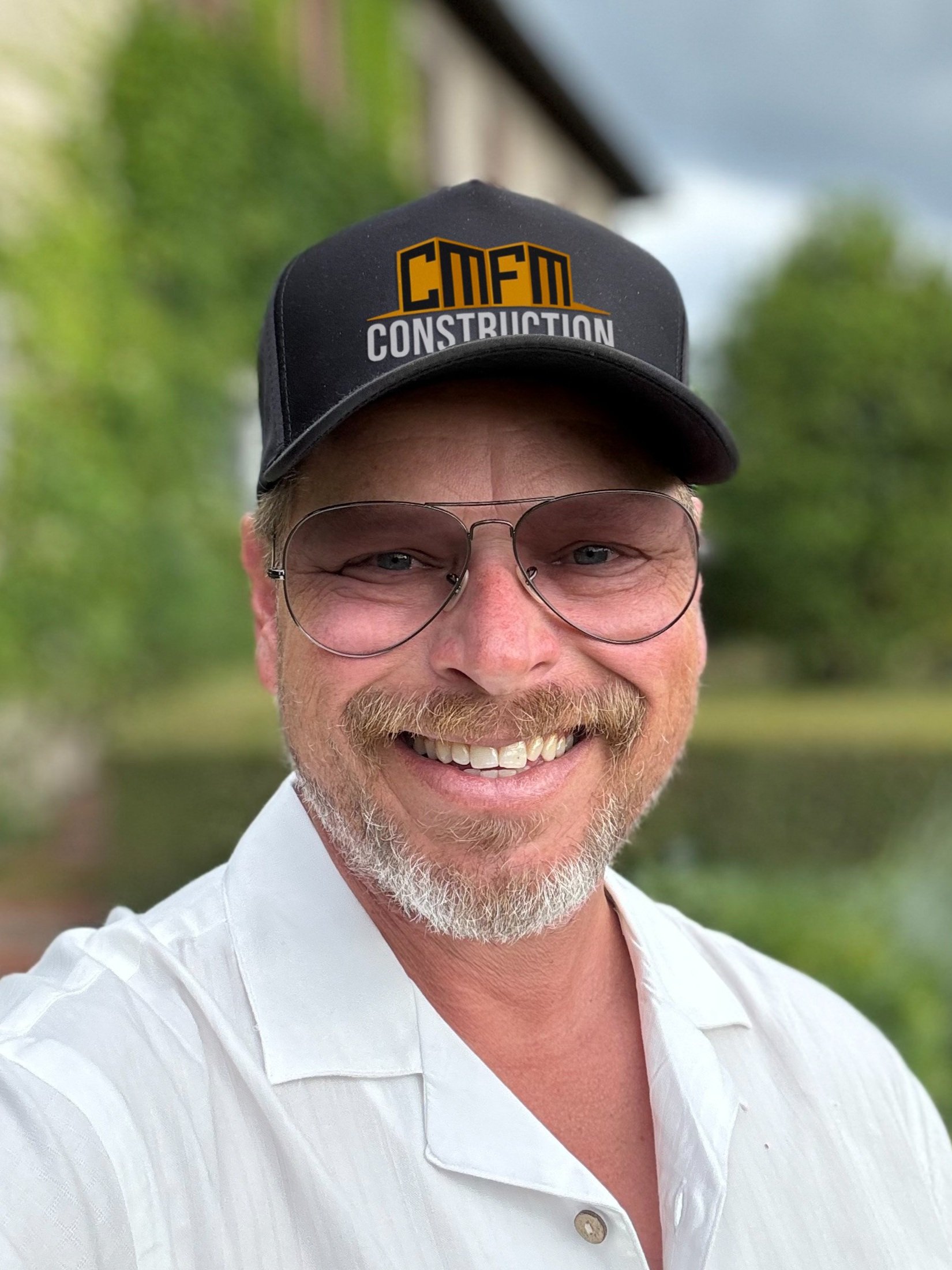 A smiling man with glasses and a beard wearing a black construction cap with the text 'CMFM Construction' and a white collared shirt, outdoors with green trees and a building in the background.
