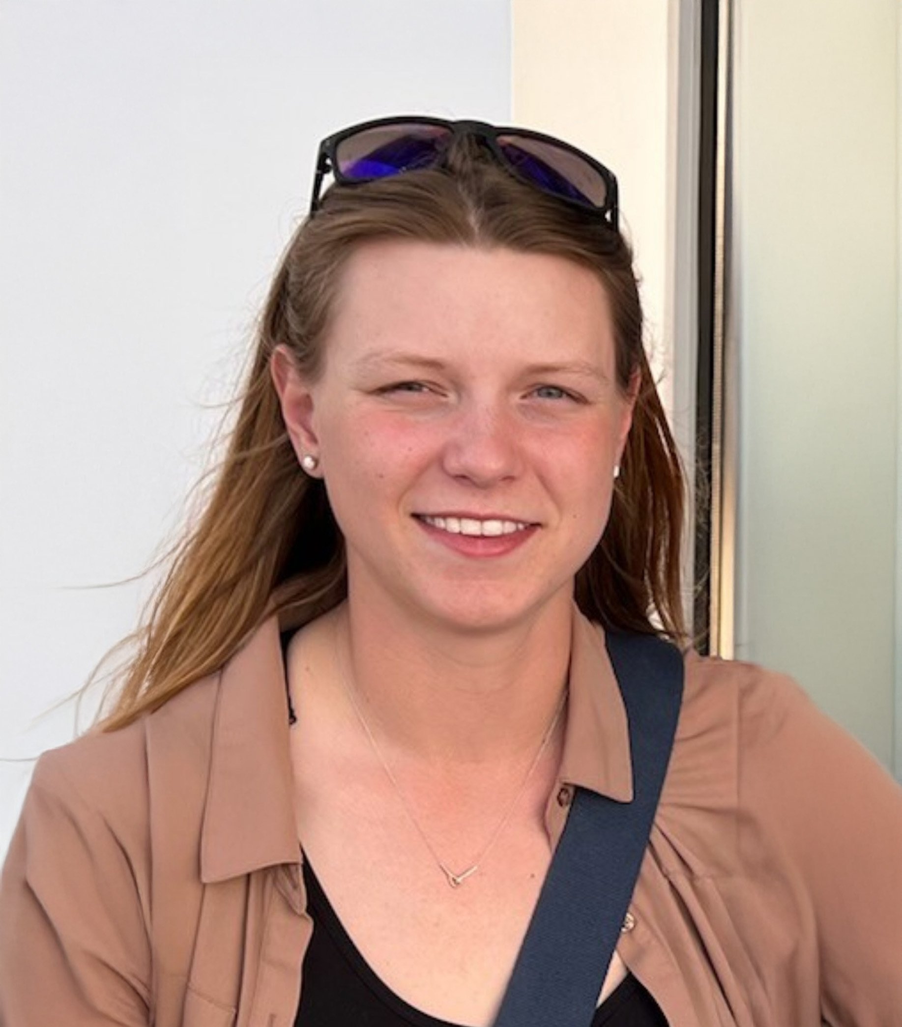 A young woman with long, red hair, wearing sunglasses on her head, a pearl earring, a necklace, a black top, and a beige jacket, smiling at the camera.