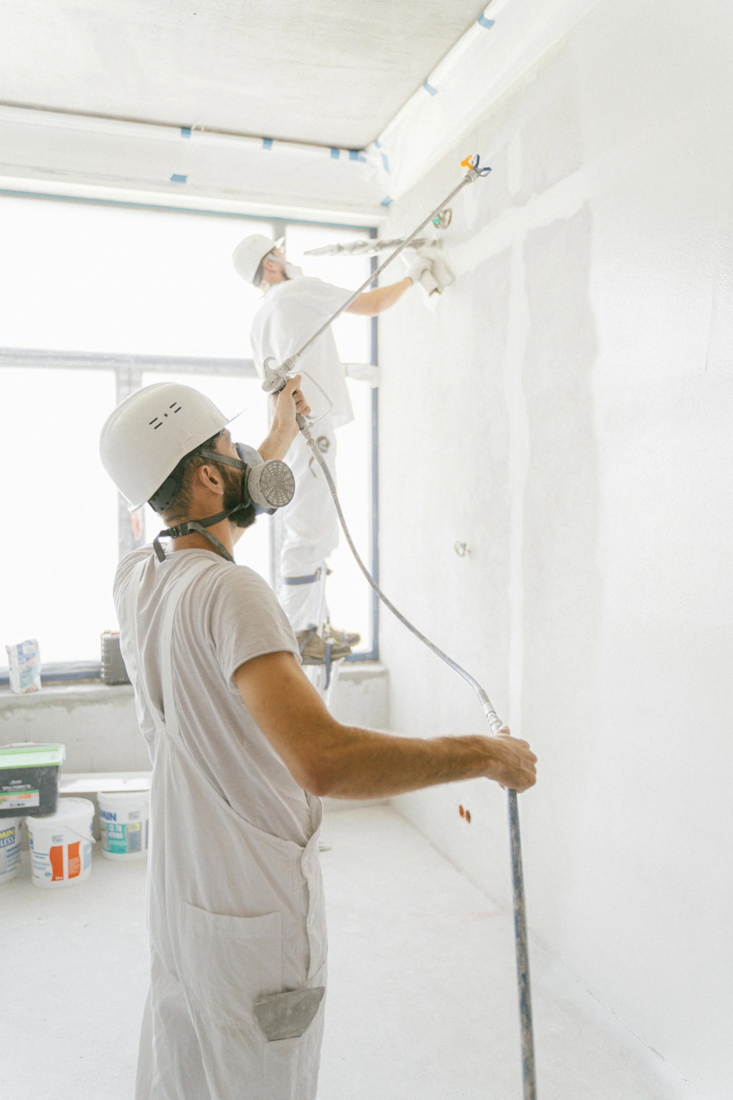 Two construction workers in white uniforms and safety gear spray insulation foam on a wall inside a building under construction.