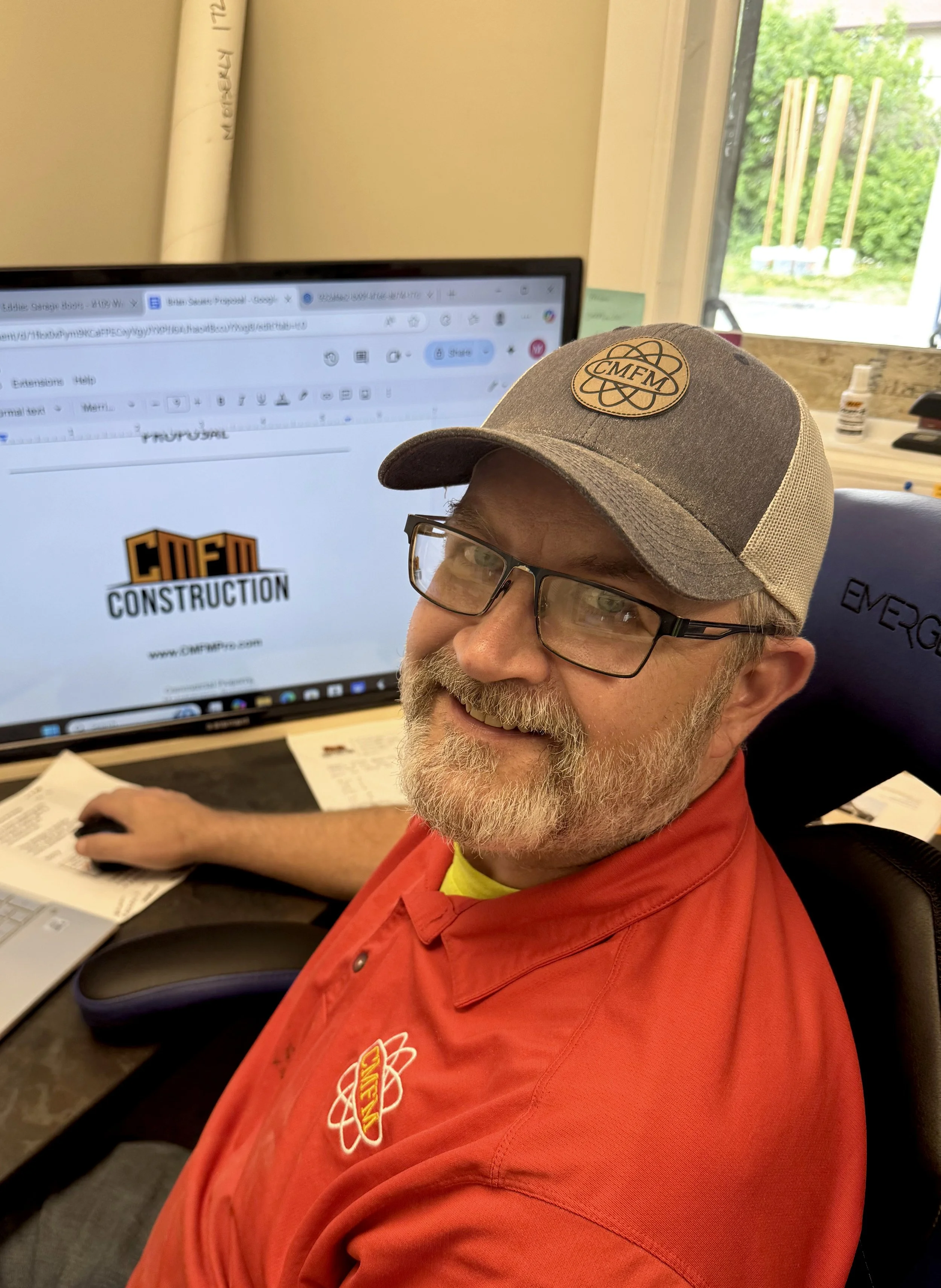 A man with glasses and a beard, smiling while sitting at a desk. He is wearing a grey and beige cap and a red shirt with an atom logo and the word 'ENERG' on it. In the background, there is a computer monitor displaying a logo that says 'CMFM Construction' and a window showing greenery outside.
