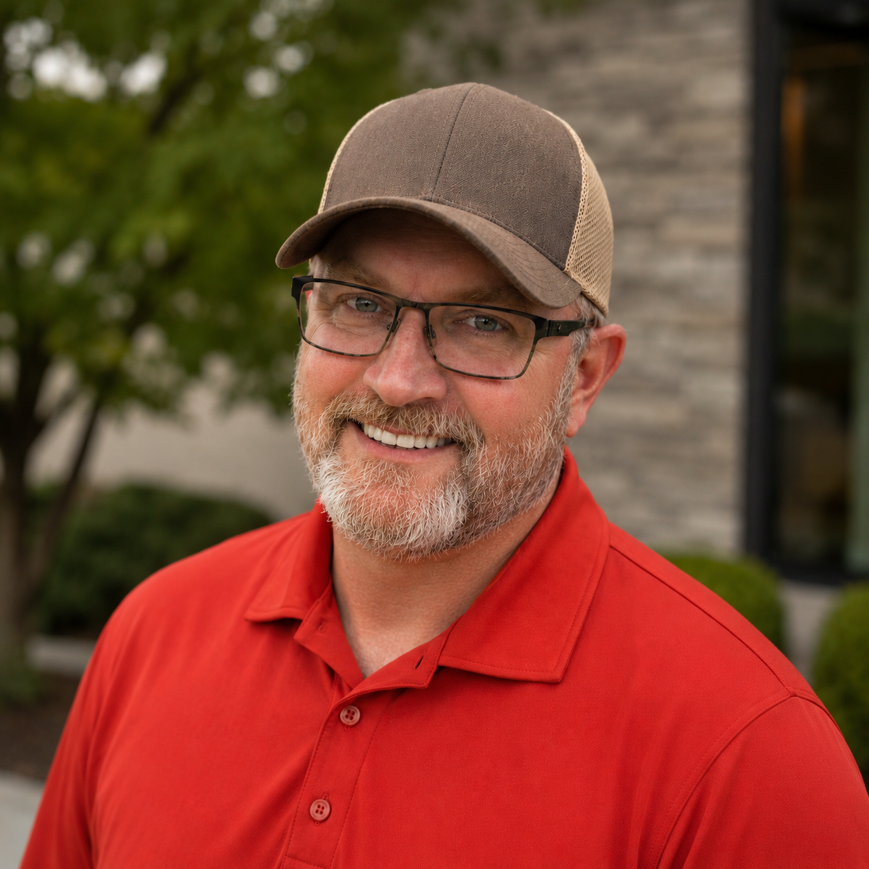 A man with glasses and a beard, smiling while sitting at a desk. He is wearing a grey and beige cap and a red shirt with an atom logo and the word 'ENERG' on it. In the background, there is a computer monitor displaying a logo that says 'CMFM Construction' and a window showing greenery outside.
