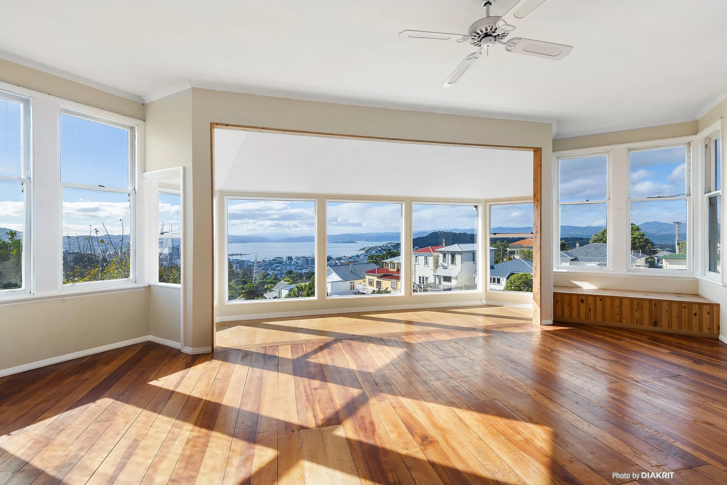 Empty living room with large windows and panoramic city and water view, hardwood flooring, ceiling fan, and sunlight.