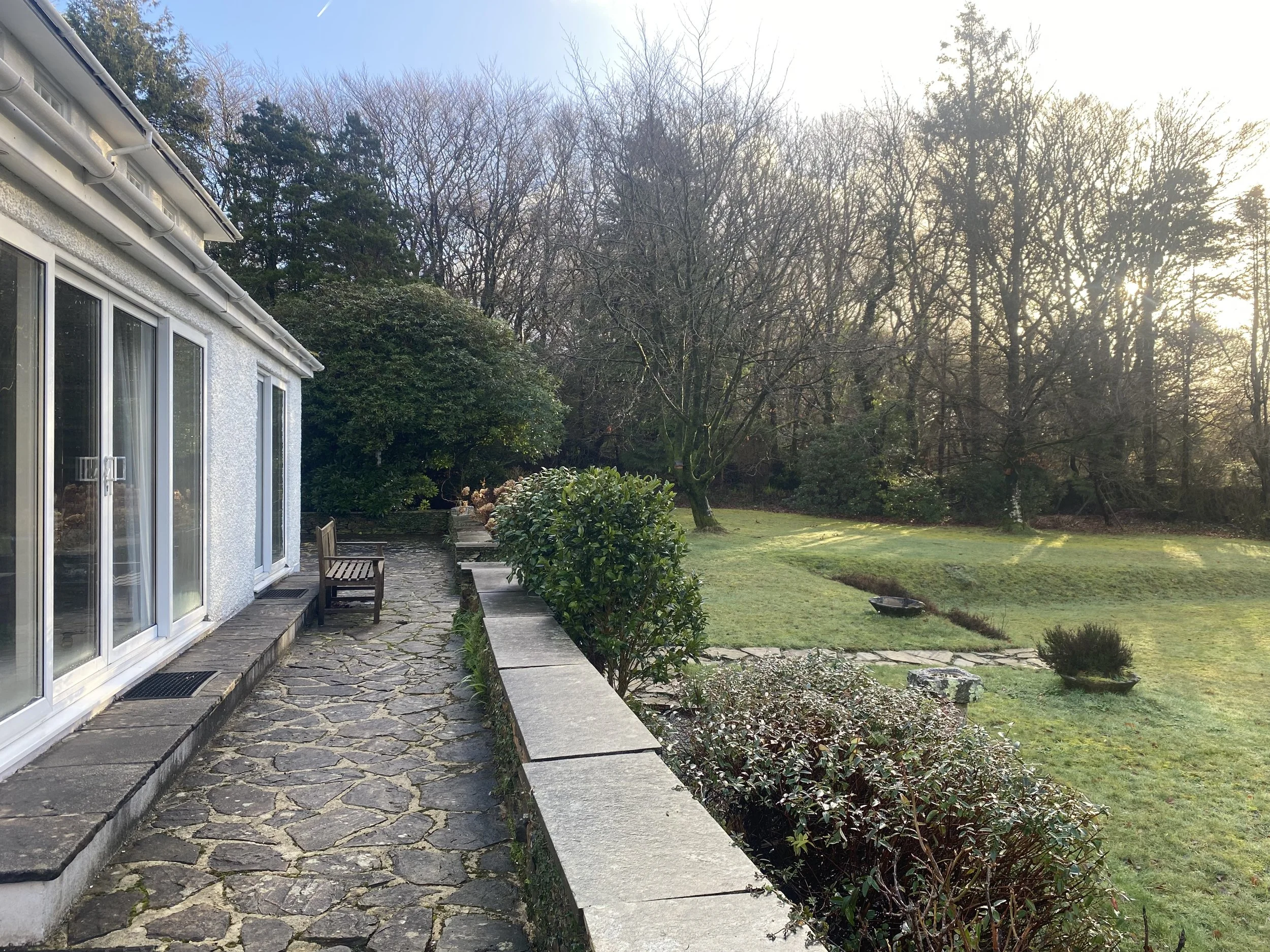 A backyard garden with a stone pathway, green bushes, and trees in the background, near a white house with large glass sliding doors.
