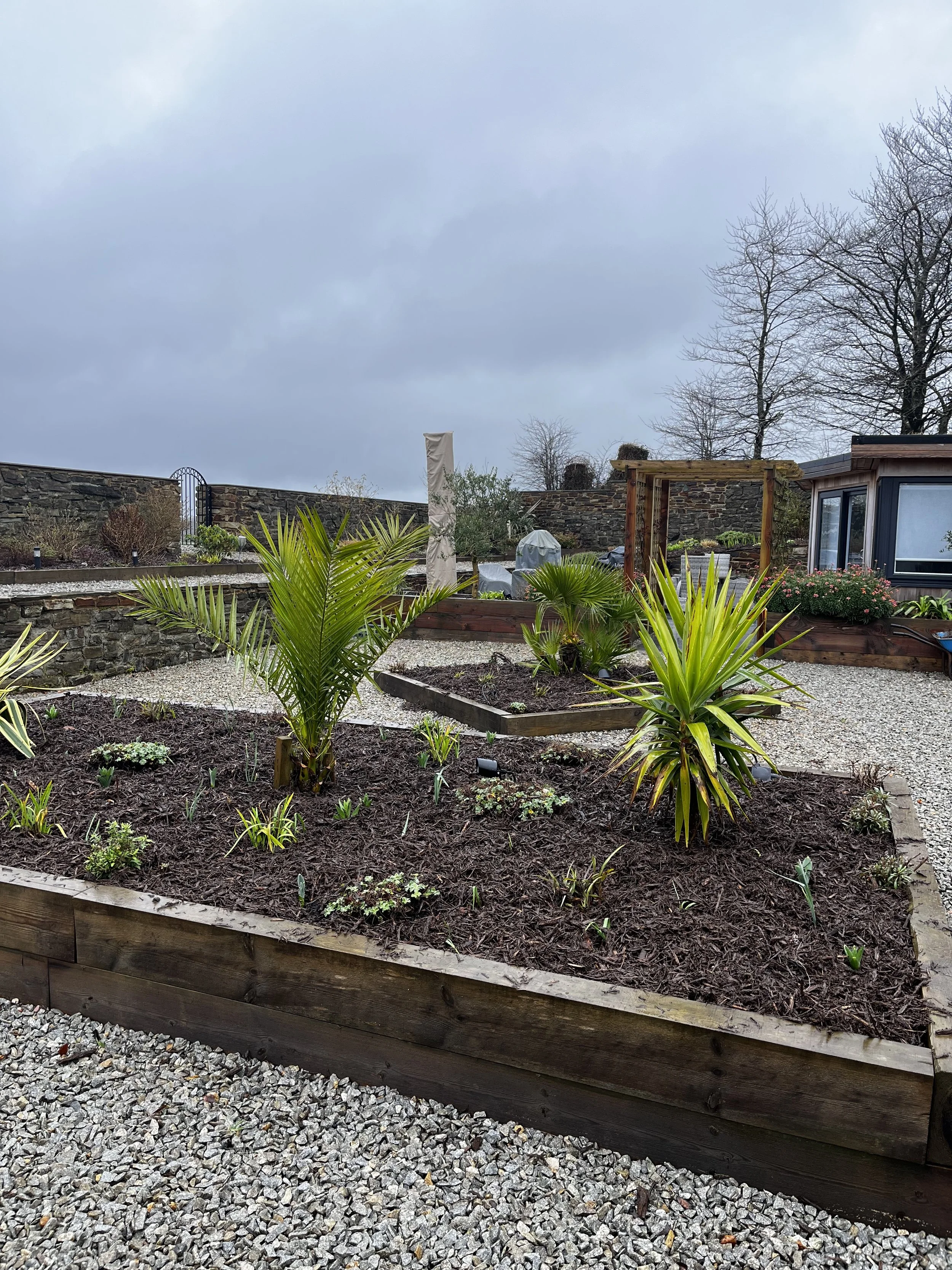 A sunken garden with raised wooden beds containing various plants and young trees, a gravel pathway, and a stone wall with a metal gate, under a cloudy sky.