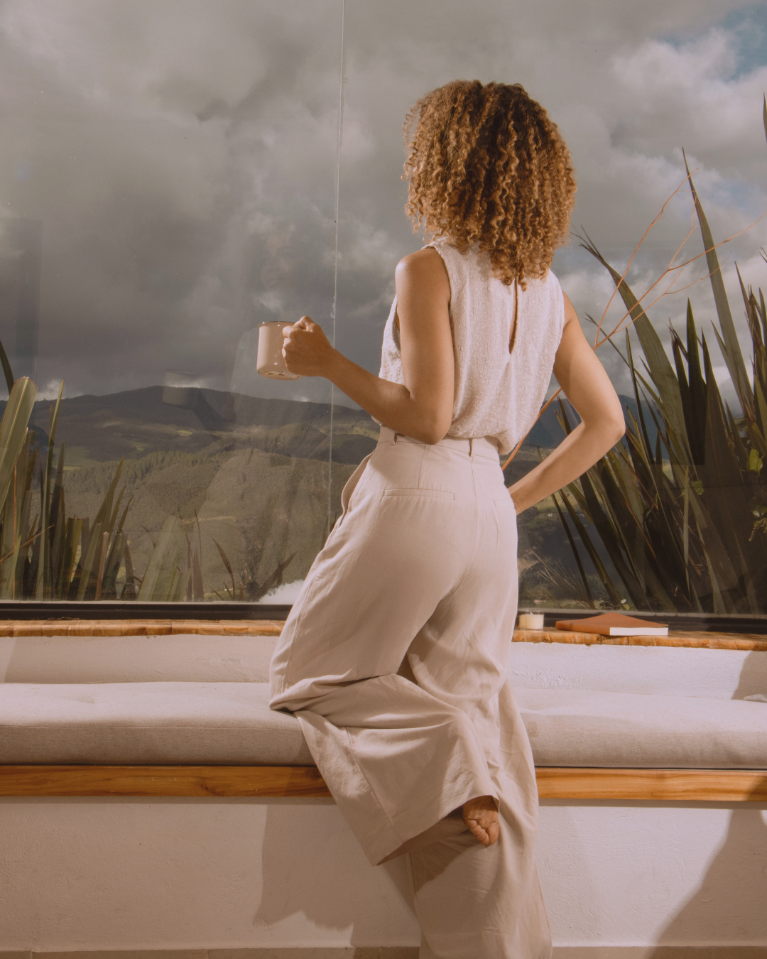Woman with curly hair sitting on a window ledge, looking outside at a scenic landscape with mountains and cloudy sky, holding a mug in her right hand.
