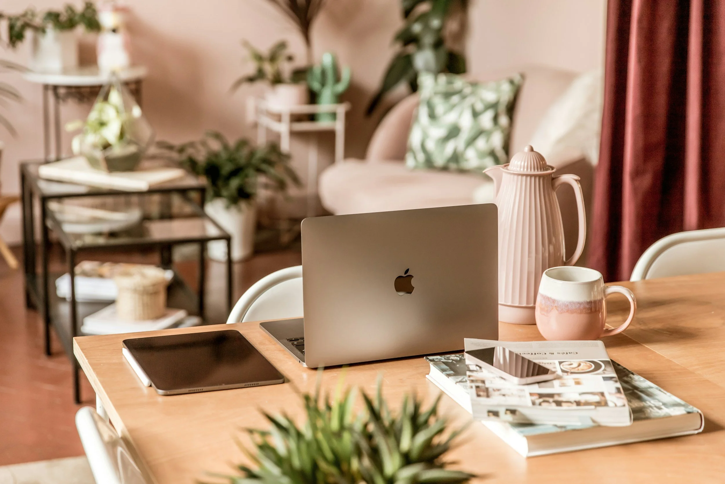 A wooden table with an Apple MacBook, a tablet, a smartphone on a stack of magazines, a pink ceramic mug, a pink and white teapot, and a potted plant in the foreground. In the background, there is a sofa with green patterned pillows, a small black metal side table with books and a plant, and a white plant stand with multiple plants. The room is decorated with pink curtains and houseplants.