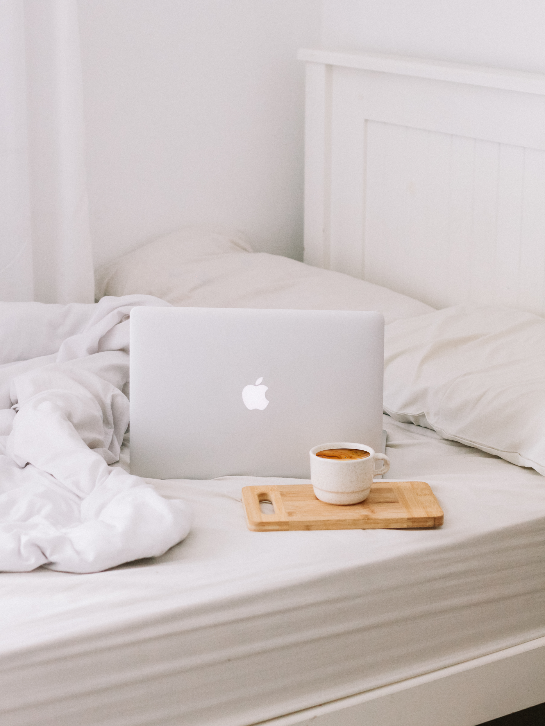 A laptop computer, a mug of coffee, and a wooden tray sit on a white bed with white bedding in a minimalistic, bright bedroom.