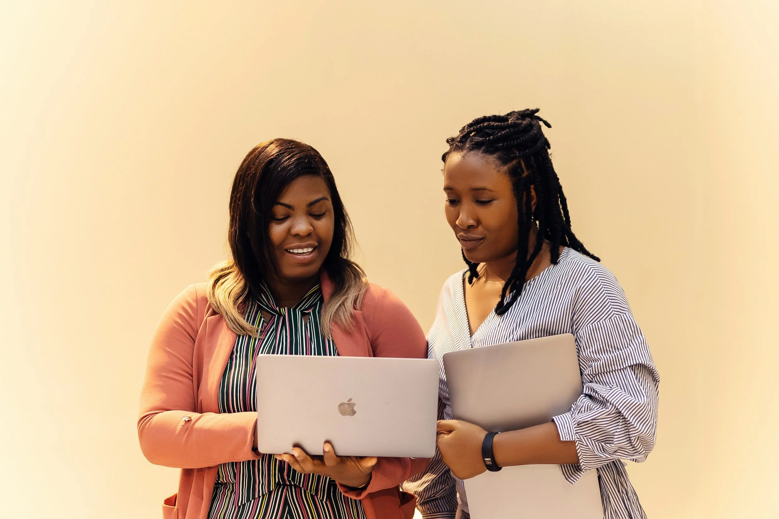 Two women looking at a laptop together in an indoor setting with a plain light-colored wall in the background.