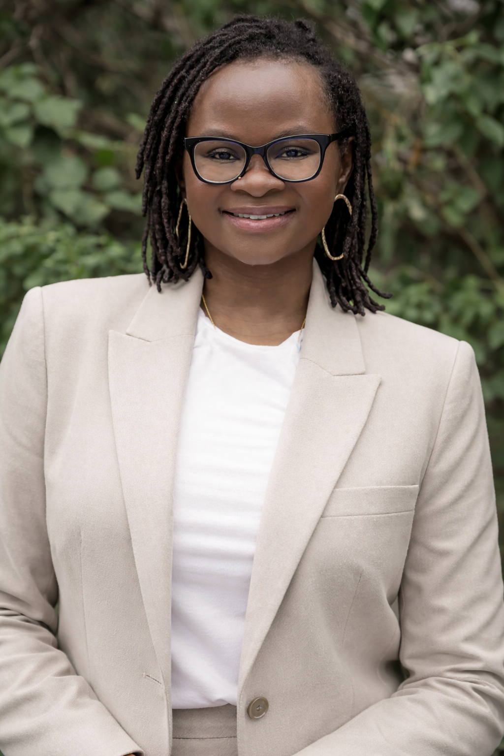 A woman with dark skin, dreadlocks, black glasses, and gold hoop earrings, smiling outdoors in front of green foliage, wearing a beige blazer and white shirt.