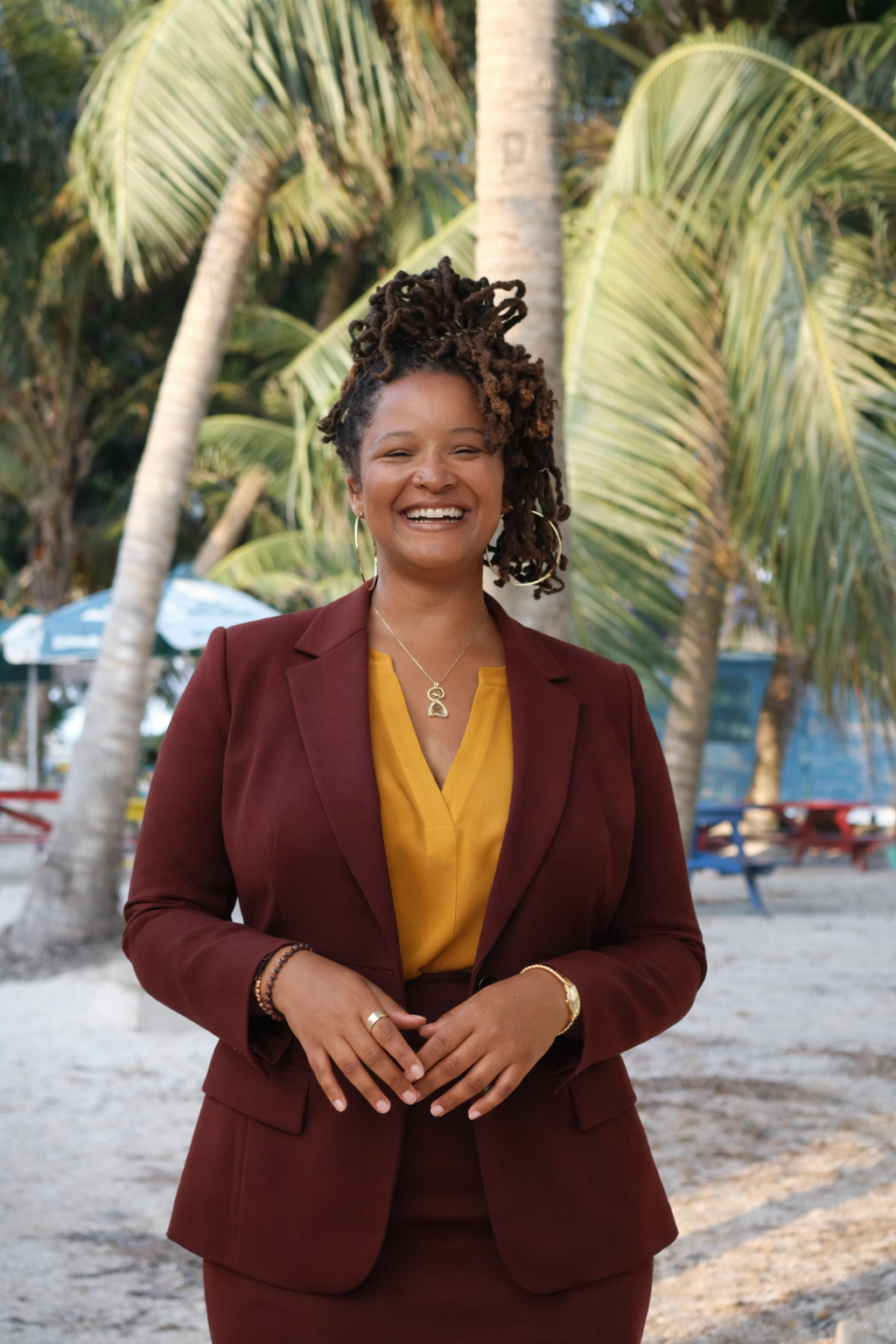 A smiling woman in a maroon blazer and yellow blouse standing outdoors with palm trees and beach scenery in the background.