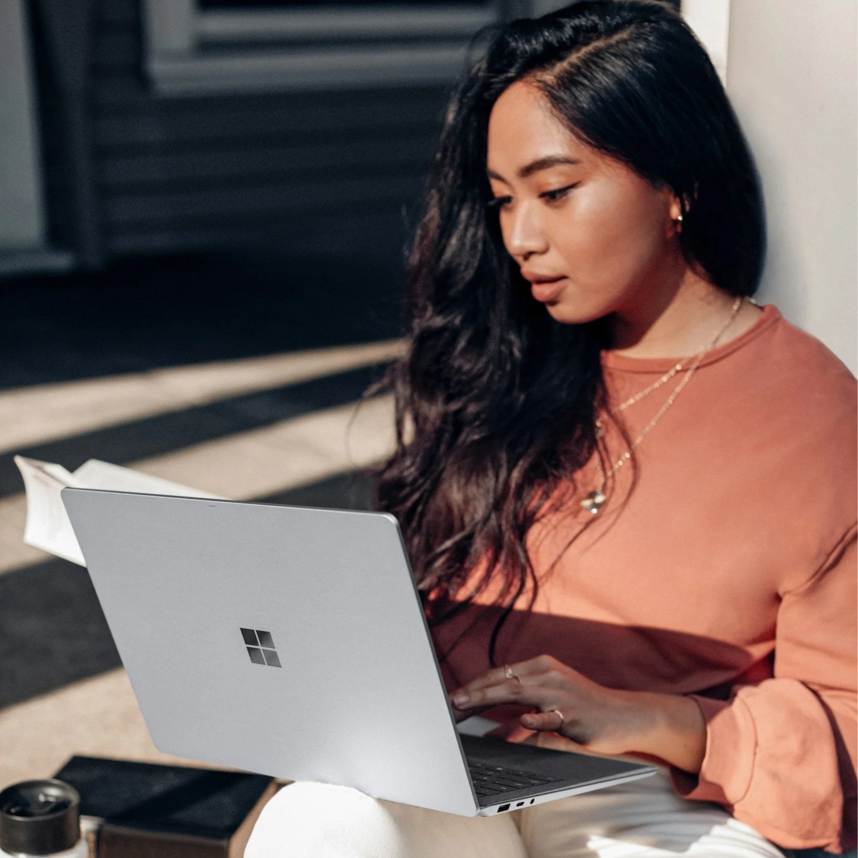 A woman with long black hair and light brown skin sitting and looking at a Microsoft Surface laptop, wearing an orange long-sleeve shirt, with her right hand on the laptop keyboard and her left hand resting on her lap, in a casual indoor setting.