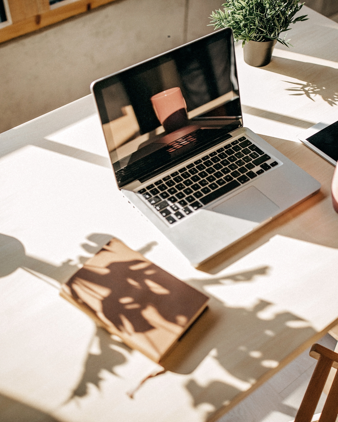 A laptop, a plant, and a closed tablet on a wooden table with sunlight casting shadows.