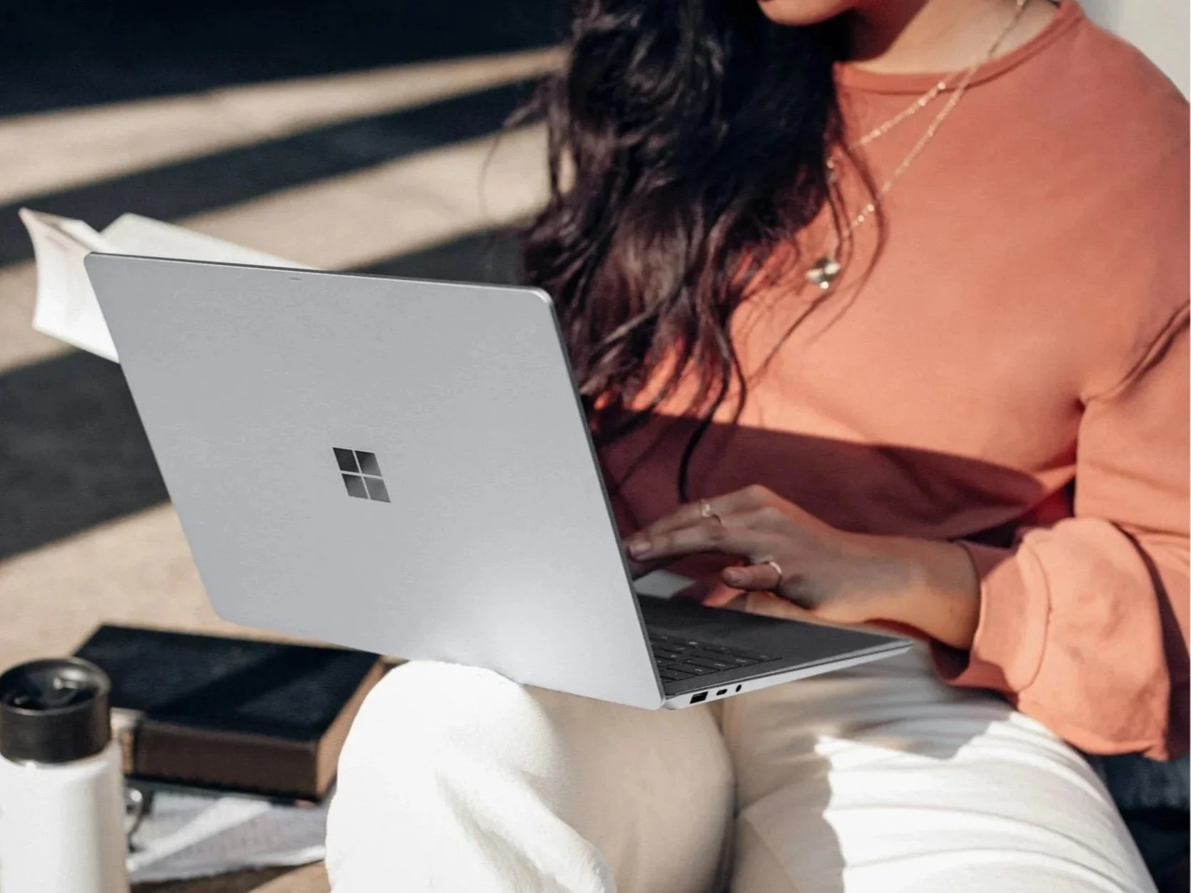 A woman with long dark hair working on a Microsoft Surface laptop, sitting at a table with a notebook, pen, and water bottle, wearing a pinkish-orange top and white pants.