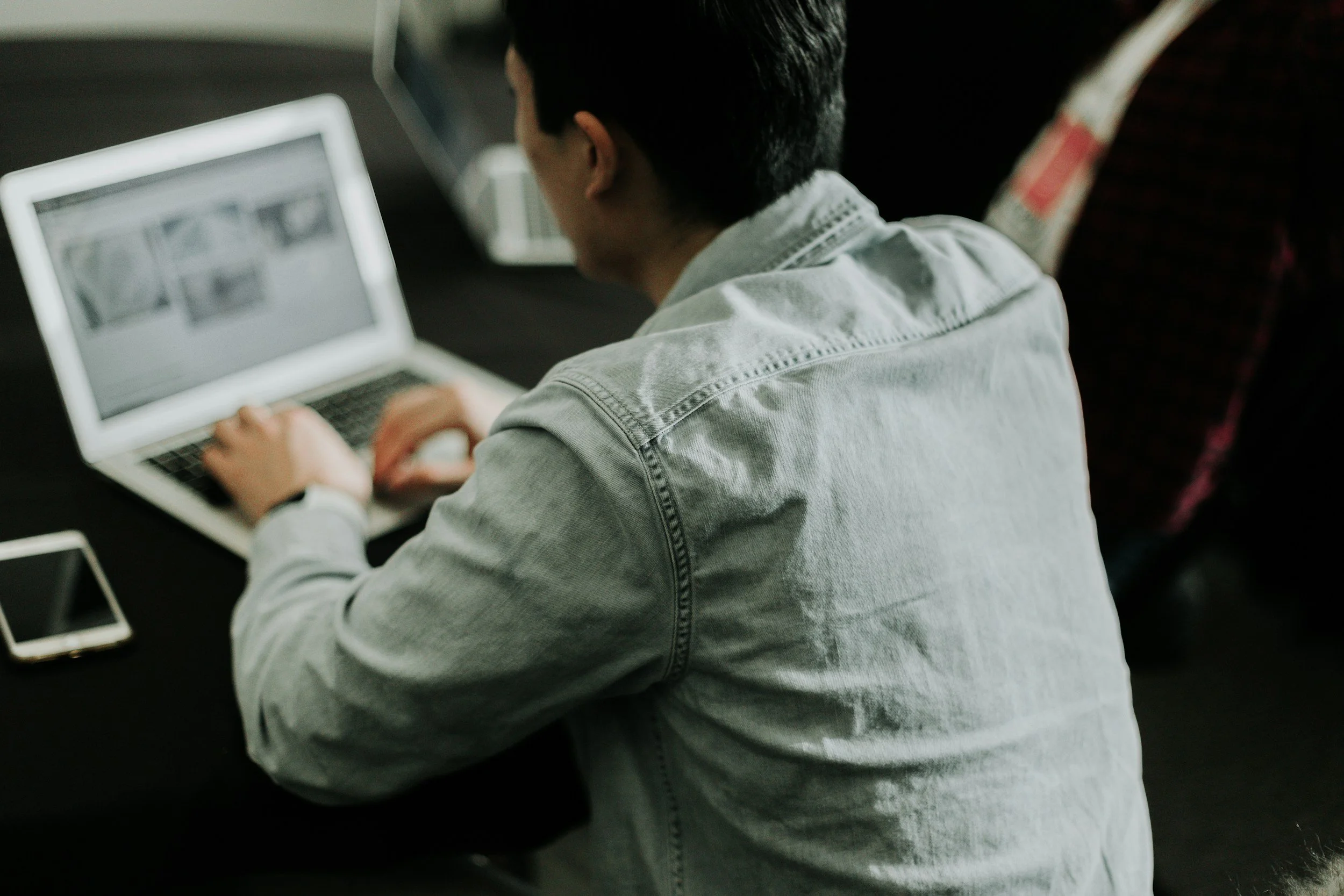 A person with dark hair wearing a light gray denim jacket sitting at a black table, working on a silver laptop, with a smartphone placed beside them.
